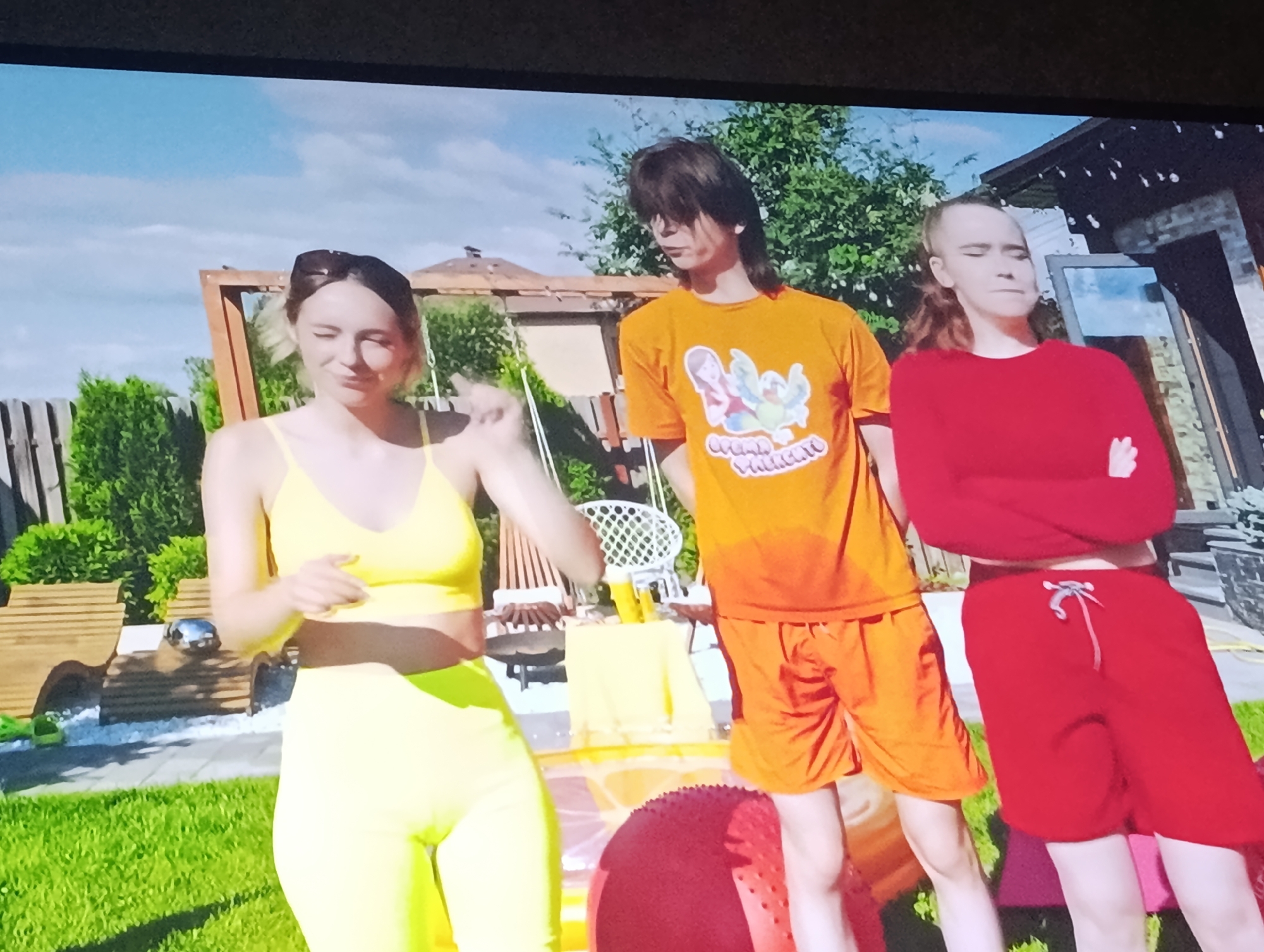 Three young people are standing in a backyard on a sunny day. The person on the left is a woman wearing a yellow sports bra and yellow leggings. She has blonde hair pulled back and is wearing sunglasses on her head. She is looking down and to her left, with her right hand extended. The person in the middle is a young man with dark, shaggy hair wearing an orange t-shirt with a colorful graphic on the front and orange shorts. He is looking forward with a neutral expression. The person on the right is a young woman with her hair pulled back and wearing a red long-sleeved shirt and red shorts. She has her arms crossed and is looking down. In the background, there is a wooden swing set, trees, a fence, and a brick house. There is also a table with yellow items on it and a large red exercise ball on the ground. There are no visible landmarks that specifically place this scene in the city of Dnipro, Ukraine.
