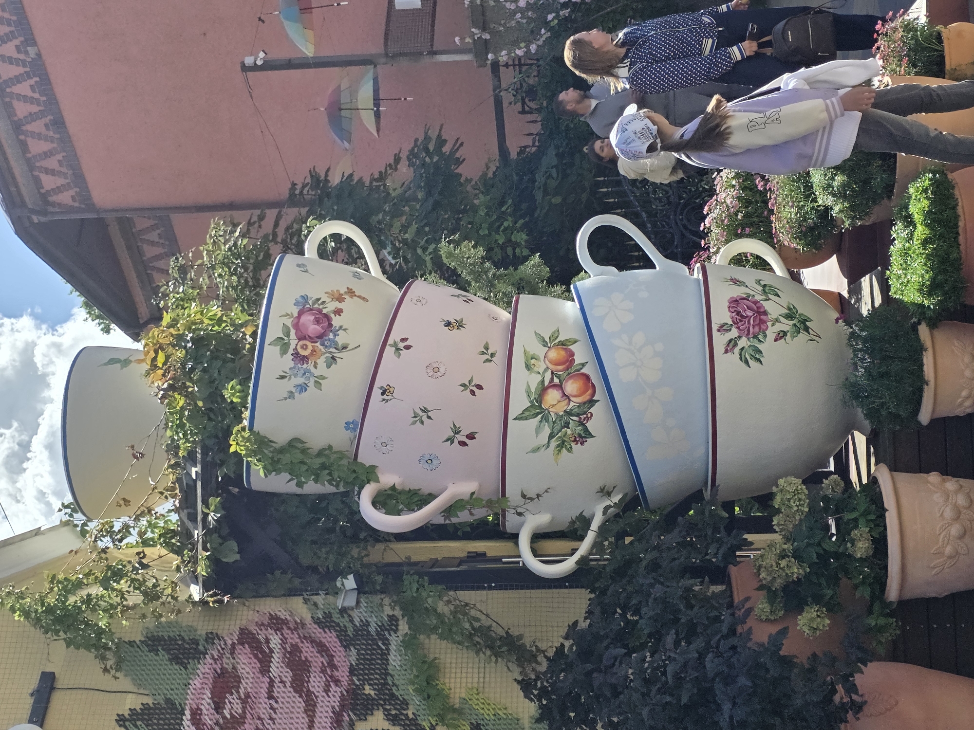 The image captures an outdoor scene in Zelenogradsk, Russia, featuring a whimsical display of oversized teacups stacked vertically. The teacups are decorated with floral patterns and fruit motifs. Green ivy vines are cascading down the sides of the teacups. In the background, a pink building with decorative brickwork is visible, along with several small, colorful umbrellas suspended in the air. A group of people are present, including a young girl wearing a baseball cap and a purple jacket, standing near the teacup display. A woman in a blue and white patterned jacket and another person, possibly a man, are also in the background, observing the scene. The foreground is occupied by potted plants, adding to the garden-like atmosphere. The overall impression is one of a charming and artistic urban setting.