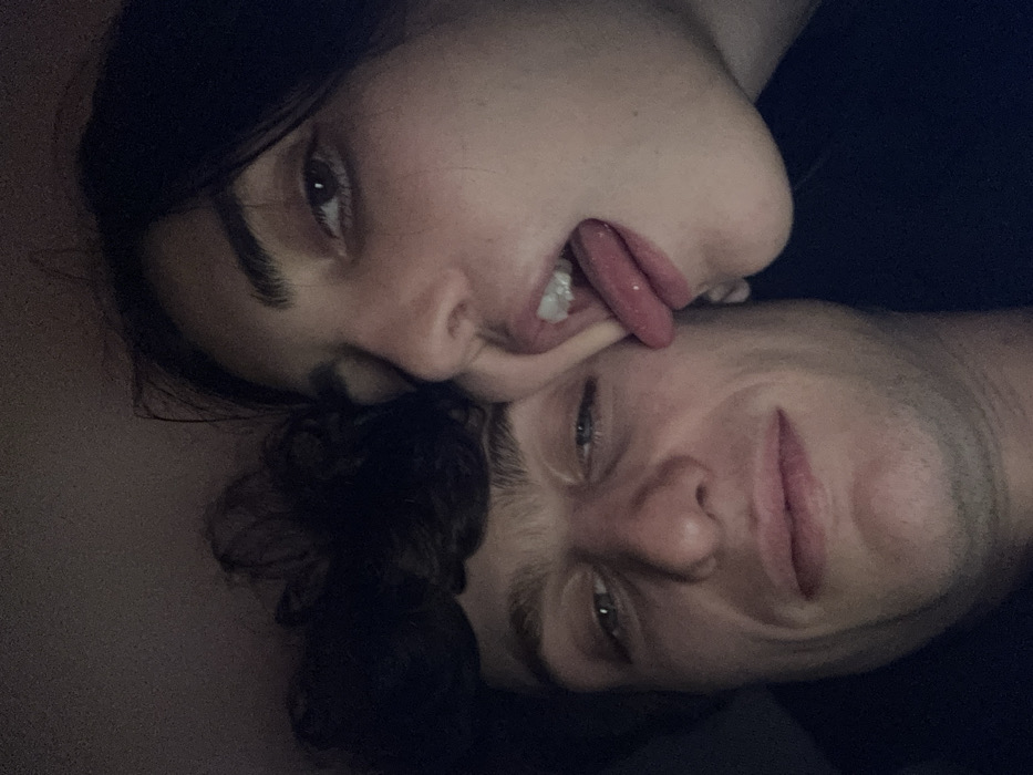 The image depicts two individuals, a male and a female, positioned in close proximity within a dark, indistinct environment. The male, with dark curly hair and light skin, is on the left, looking directly towards the viewer with a slight smile and his left eye partially closed. The female, also with dark hair and light skin, is positioned to the right of the male, her face partially visible. Her mouth is open with her tongue extended forward, appearing to make contact with the male's lower cheek or jawline. Her right hand, with visible fingers, is pressing against her own cheek near her mouth, creating a distortion of her facial features. The scene is illuminated by dim, possibly artificial, lighting. The specific location in the city of Paris, France, cannot be determined from the visual information provided.