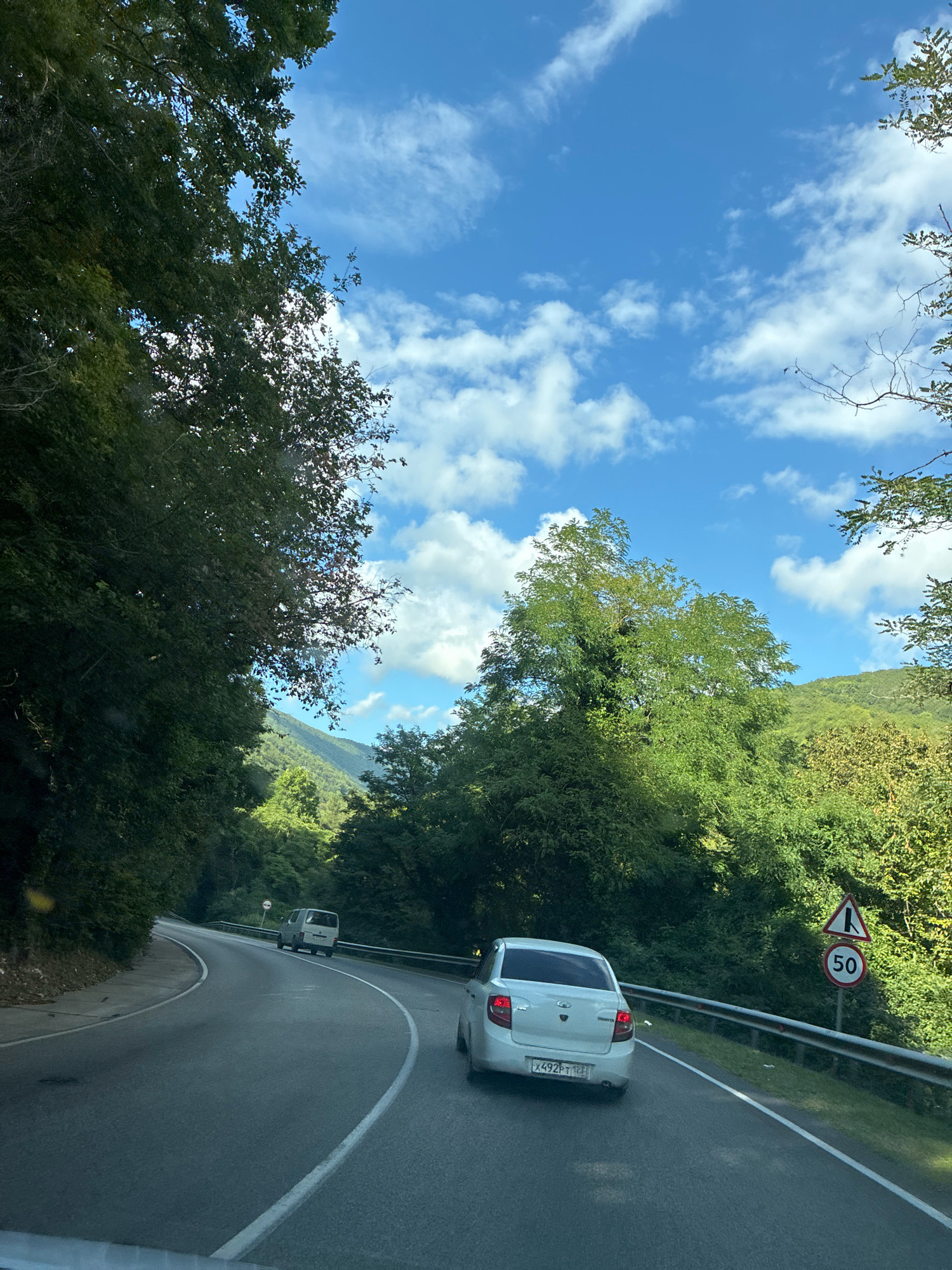 The image depicts a winding mountain road with a clear blue sky filled with fluffy white clouds. Lush green trees line both sides of the road, with hills and mountains visible in the distance. A white sedan with a license plate reading "X492PT 123 RUS" is in the foreground, followed by a white van further up the road. A speed limit sign indicating 50 is visible on the right side of the road, along with a warning sign for a sharp turn. The image appears to be taken from a vehicle driving on the road. The scene is consistent with the mountainous regions surrounding Sochi, Russia, likely on a road leading into or out of the city.