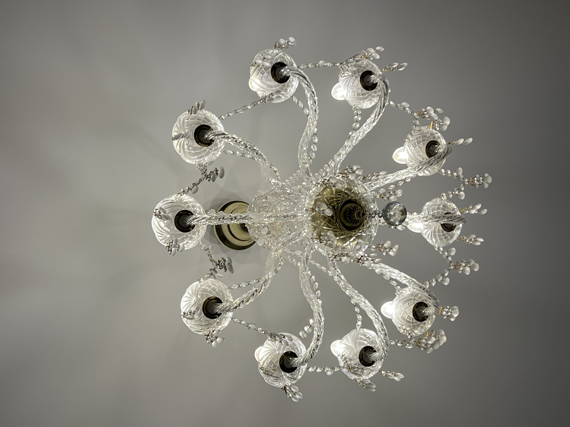A close-up, eye-level shot showcases an ornate crystal chandelier hanging from a plain, light gray ceiling. The chandelier features multiple arms, each holding a clear, intricately carved glass shade. Delicate crystal beads and strands hang from the arms, refracting light. The central part of the chandelier has a brass-colored fixture with detailed engravings. The image is well-lit, with the light emanating from the chandelier itself. No people or other objects are visible. The context of Veliky Novgorod, Russia is not discernible from the image itself.