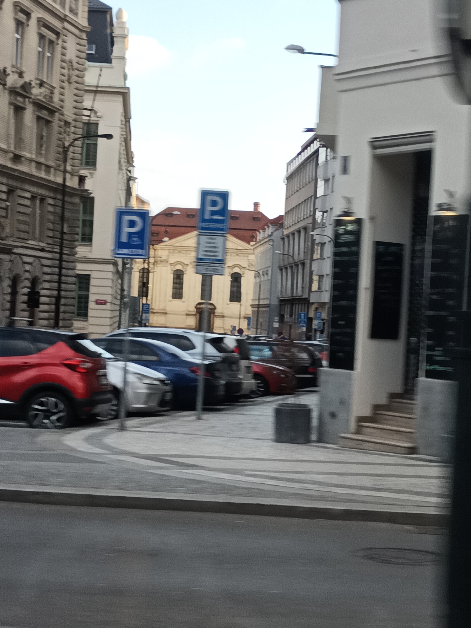 The image depicts a street scene in Prague, Czechia. In the foreground, a gray asphalt road is visible, bordered by a raised concrete curb. Beyond the curb is a pedestrian walkway paved with gray and white interlocking stones. A row of parked cars lines the walkway, including a red SUV, a silver sedan, a blue sedan, a dark SUV, and a dark red sedan. Parking signs with a blue "P" symbol are posted along the street.
The background features a mix of architectural styles. On the left, a building with ornate stone facades and arched windows suggests historical architecture. Adjacent to it is a modern building with multiple floors and rectangular windows. Further down the street, a light yellow building with a red tiled roof and arched windows appears to be a significant structure, possibly a church or public building. On the right side of the image, a white building with a modern design has an entrance with stairs leading up to it. Black boards displaying text, possibly menus or advertisements, are mounted on this building. The sky is overcast. The overall impression is a busy urban environment with a blend of old and new buildings.