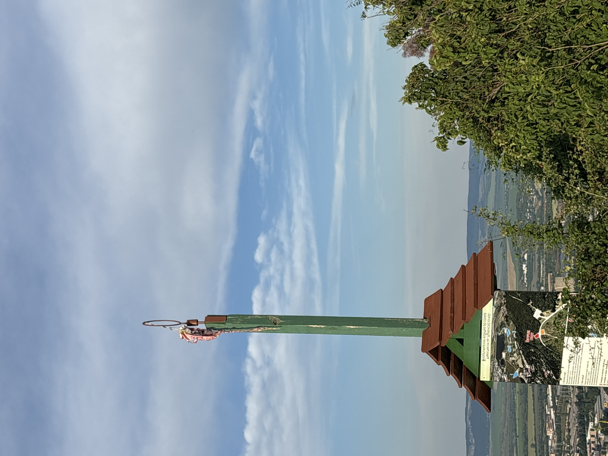 This is an eye-level, medium outdoor shot of a tall, green wooden pole topped with a metal ring from which pink and white fabric scraps are hanging. The pole is mounted on a small, red-roofed structure, which is affixed to a signpost. The sign displays a map of the area with various points of interest highlighted, and text in a foreign language. The background consists of a hazy, distant landscape of rolling green hills, fields, and a town. Lush green foliage is visible in the foreground, partially obscuring the signpost and the town below. The sky is a bright blue with scattered white clouds. The scene appears to be a scenic overlook or a trail marker.