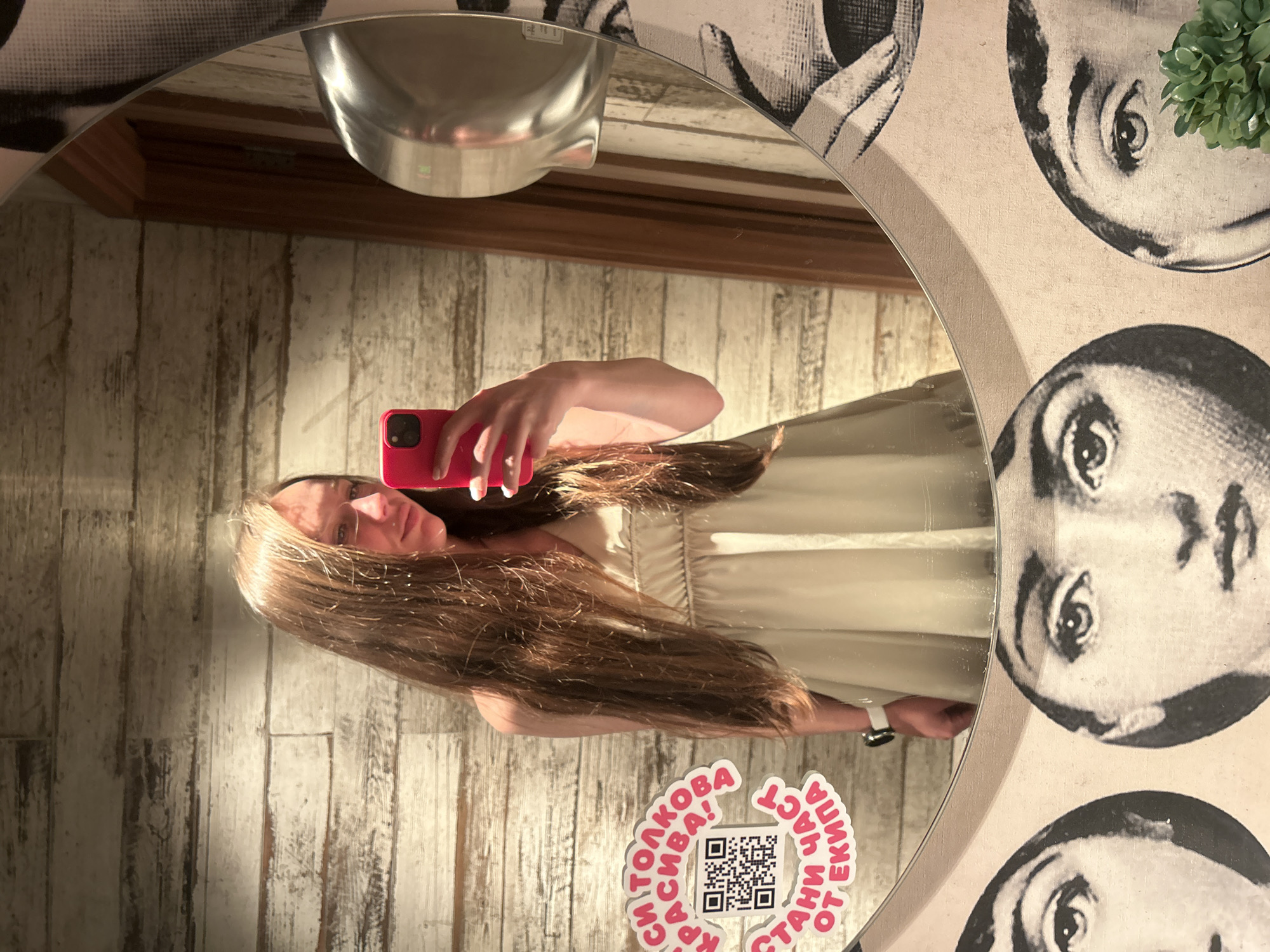 A female is captured in a mirror selfie within an interior setting, likely a public restroom, in Sofia, Bulgaria. She has long, straight, light-brown hair, visible cascading over her shoulders and chest. She wears a light beige sleeveless dress with a V-neckline. Her right hand, holding a red-cased smartphone, is positioned to take the photograph, with her gaze directed towards the phone screen. A white smartwatch is present on her left wrist. The circular mirror features a decorative border adorned with repeating black and white stylized illustrations of a woman's face. Reflected behind her is a wall constructed from horizontally placed light-colored wooden planks. A silver wall-mounted hand dryer is visible in the reflection on the right side of the mirror, alongside a darker wooden door frame. A sticker on the wooden wall, partially obscured by the mirror's edge, contains a QR code and Cyrillic text that translates to "You are so beautiful! Become part of the team!" A small cluster of green artificial foliage is visible at the bottom right of the mirror's frame.