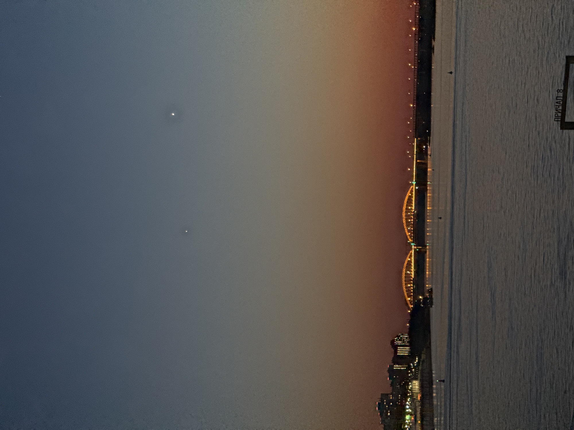 The image depicts a twilight scene over a river in Nizhny Novgorod, Russia, showcasing the Nizhny Novgorod Metro Bridge across the Oka River. The sky presents a gradient transition from dark blue at the top to orange and red near the horizon, with visible grain/noise throughout. Two distinct, bright white points of light are visible in the upper sky, along with a fainter white point near the top right corner. The lower half of the image is dominated by a dark, calm body of water (the Oka River), with two dark conical buoys floating on its surface. A small, dark vessel is situated on the water between the left bank and the bridge.
On the left bank, a collection of multi-story buildings with numerous illuminated windows is visible. A large bridge with two prominent, golden-lit arched sections spans the river horizontally across the mid-ground, its deck also illuminated by a series of smaller lights. In the bottom right foreground, a dark rectangular sign partially emerges from the water, displaying white Cyrillic text that reads "ПРИЧАЛ 8" (PRICHAL 8), meaning "Pier 8" or "Berth 8." No human figures are discernible in the scene. The lighting is a blend of natural twilight and artificial illumination from the city buildings and the bridge.