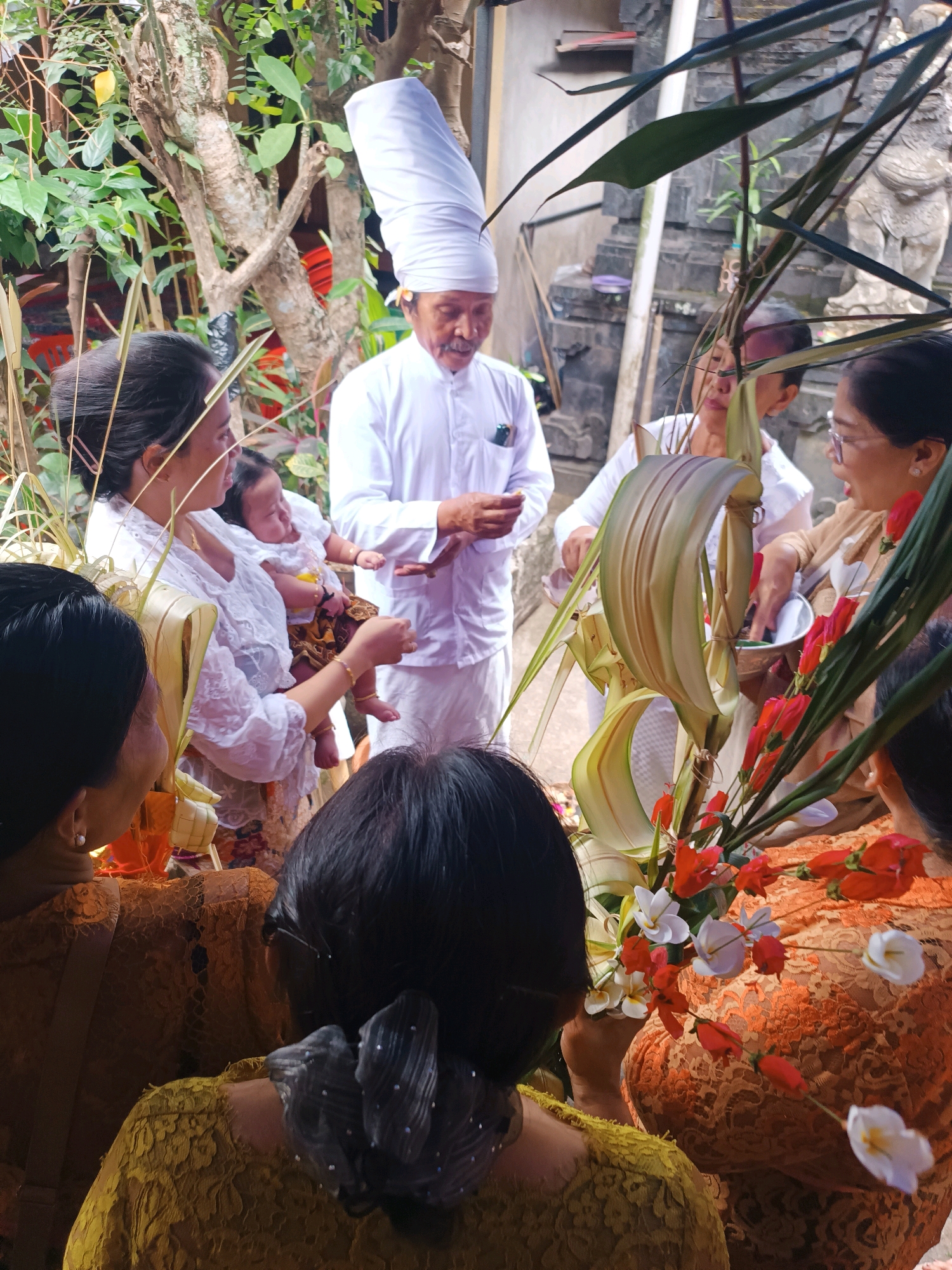 This image, taken in Tabanan, Indonesia, depicts a ceremonial gathering outdoors, likely in a temple courtyard given the stone architecture visible in the background. Several individuals are present, all appearing to be engaged in a ritual.
In the foreground, two women are partially visible from behind, their heads and upper torsos showing. One woman wears a yellow lace top, and the other a patterned orange garment. In the center of the frame, a woman in a white lace top holds a baby, who is dressed in a patterned sarong and white top. The baby appears to be looking up and smiling.
To the left, another woman, also in a white lace top, is seen from the side, looking towards the baby. Further back and slightly to the right, a man in a white traditional outfit and a tall, conical white headdress is the focal point of the group. He is holding his hands out and appears to be speaking or performing a rite. Behind him, other individuals in white attire are visible, suggesting a formal occasion.
A prominent arrangement of greenery and red and white flowers sits in the middle right of the image, partially obscuring some of the participants. The environment is lush with foliage, and the background reveals stone structures with intricate carvings, typical of Balinese temples. The lighting suggests it is daytime, with natural light illuminating the scene. The overall atmosphere appears to be one of solemnity and community participation in a significant event.