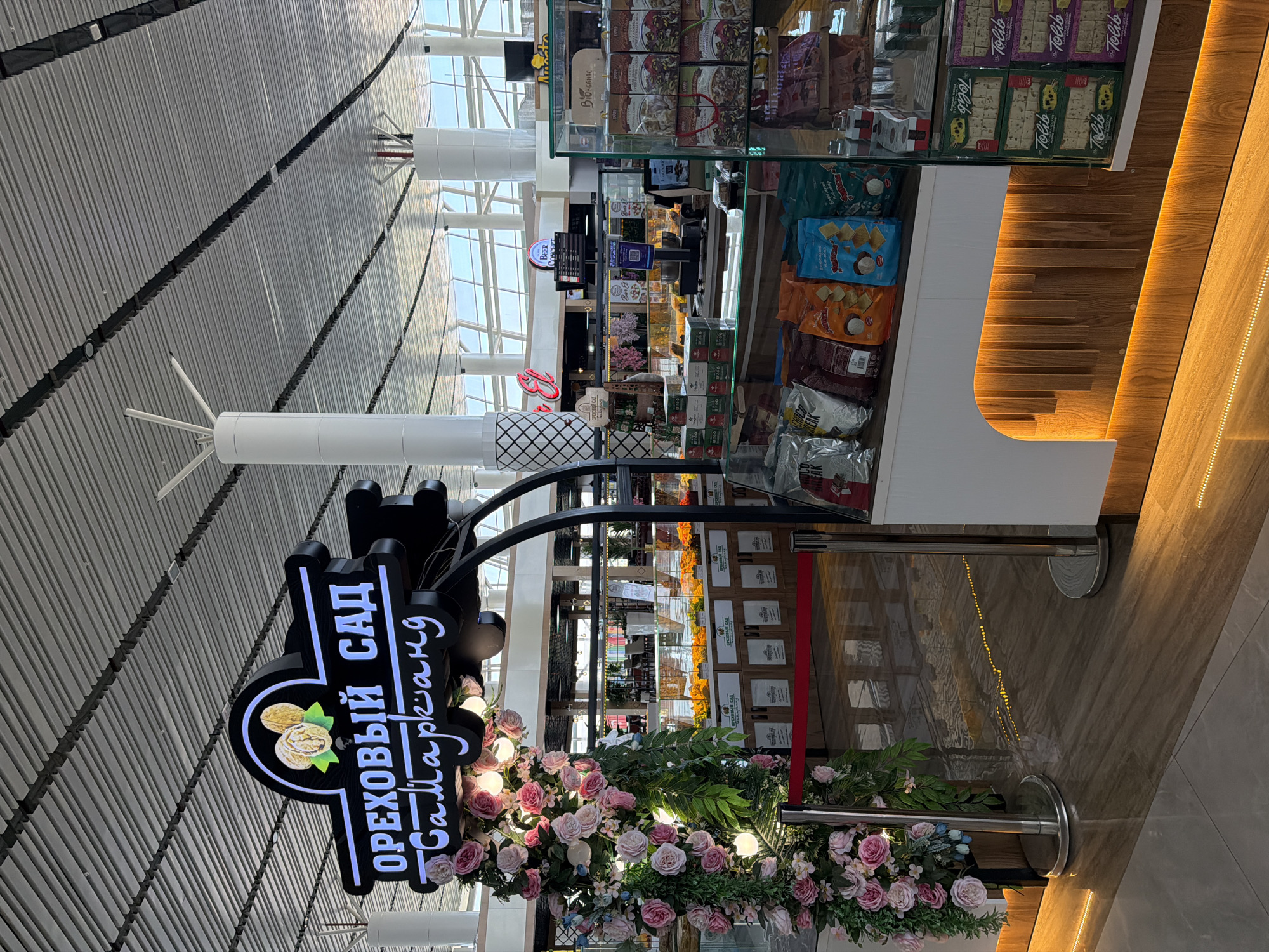 The image shows a retail stall in an indoor marketplace, possibly an airport or a large shopping center, featuring products like nuts, dried fruits, and snacks. The stall has a prominent illuminated sign in Russian that reads "ОРЕХОВЫЙ САД" (Nut Garden) and "Samarkand" written underneath. The stall is decorated with a large floral arrangement of pink and white roses and has display shelves filled with packaged goods. In the background, other stalls and shops are visible, as are seating areas and architectural elements of the building. The overall scene suggests a commercial setting.