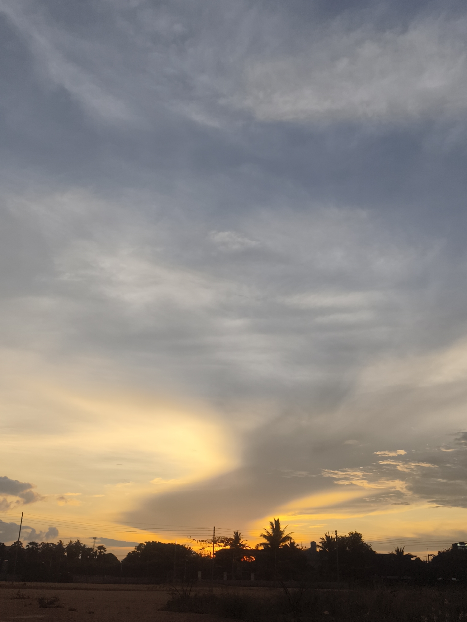The image depicts a sunset in Baan Tai, Thailand. The sky is mostly covered with clouds, transitioning from grey in the upper portion to a vibrant yellow near the horizon. A concentrated area of bright yellow light suggests the sun's position behind the trees. Below the sky, there's a silhouette of trees and vegetation. A patch of dry land is visible in the foreground. Electrical lines are strung across the scene.