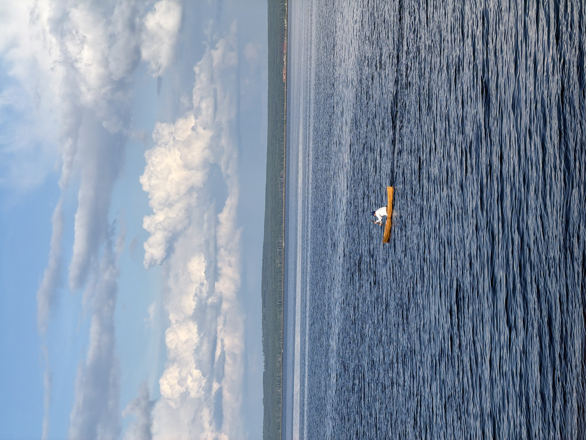 This image, when correctly oriented, captures a tranquil outdoor scene featuring a single individual on a vast body of water under a partly cloudy sky. A person, wearing a white shirt and dark trousers or shorts, is seated in a small, light brown wooden boat or canoe. The boat appears to be stationary or drifting slowly on the calm, blue water, which exhibits a finely rippled surface.
In the background, a low, dense treeline stretches across the horizon, marking the distant shoreline of a forested landmass. Above this, the sky is a clear blue, adorned with scattered white cumulus clouds of varying sizes and shapes, suggesting fair weather. The scene is illuminated by daylight, likely late morning or afternoon.
The atmosphere conveyed is one of peace and solitude, with the individual seemingly enjoying the quiet expansiveness of the water. No specific activity such as fishing is explicitly shown. There is no visible text in the image. The location context indicates this scene is near Chaikovsky, Russia, likely on a large lake or reservoir such as the Votkinsk Reservoir.