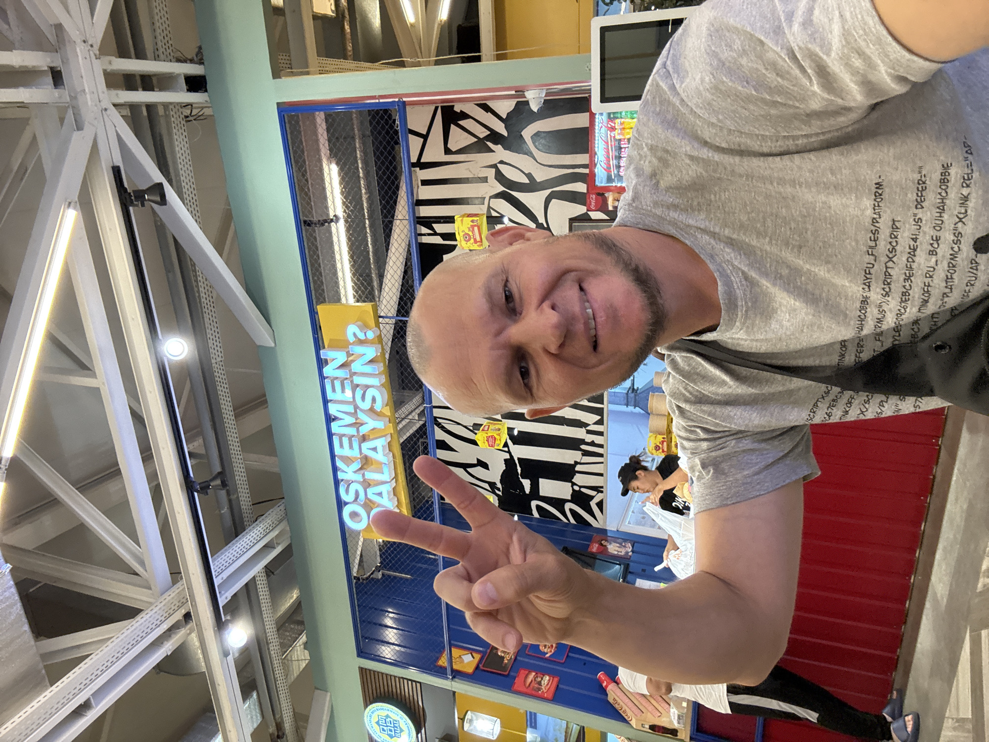 In an indoor food court, a bald, fair-skinned man with a short beard smiles at the camera, holding up two fingers in a peace sign. He is wearing a gray t-shirt with printed text and a black strap across his chest. In the background, a woman wearing a black cap and t-shirt is visible at a counter, as are shelves stocked with food items and a Coca-Cola refrigerator. Above the counter, a neon sign reads "OSKEMEN MALAYSIN?". The ceiling features industrial metal beams and lighting fixtures. The overall environment suggests a casual dining establishment in Oskemen, Kazakhstan.