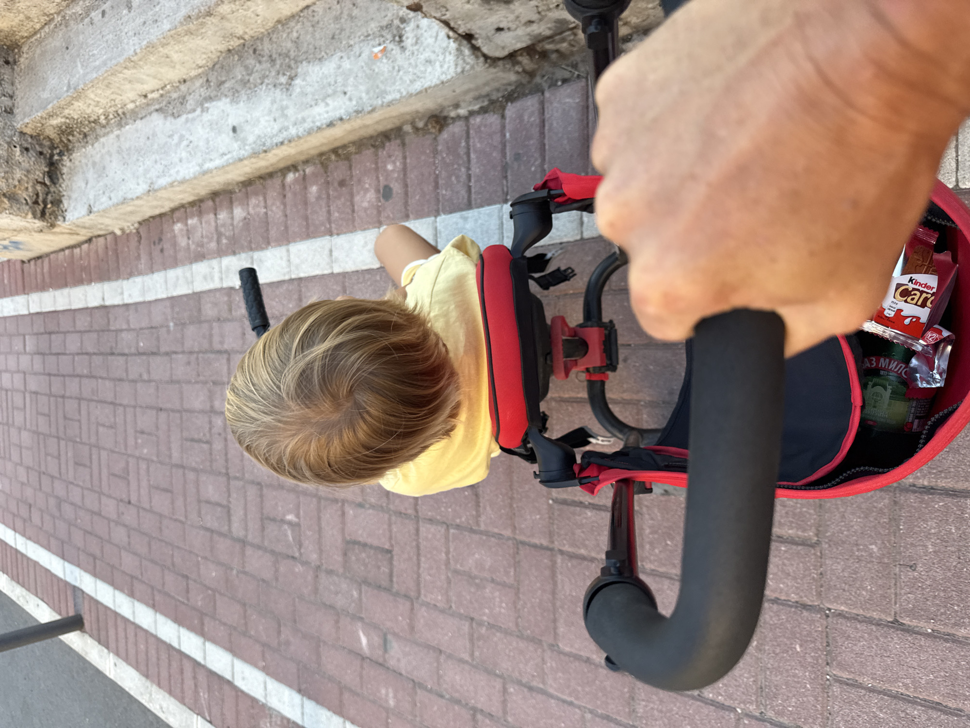The image shows a person's hand holding the handle of a red stroller, which contains a young child with blonde hair wearing a yellow shirt. The stroller is on a red brick pathway next to a concrete staircase. The background shows more of the brick pathway and a sidewalk. The scene appears to be an outdoor setting in a city.
