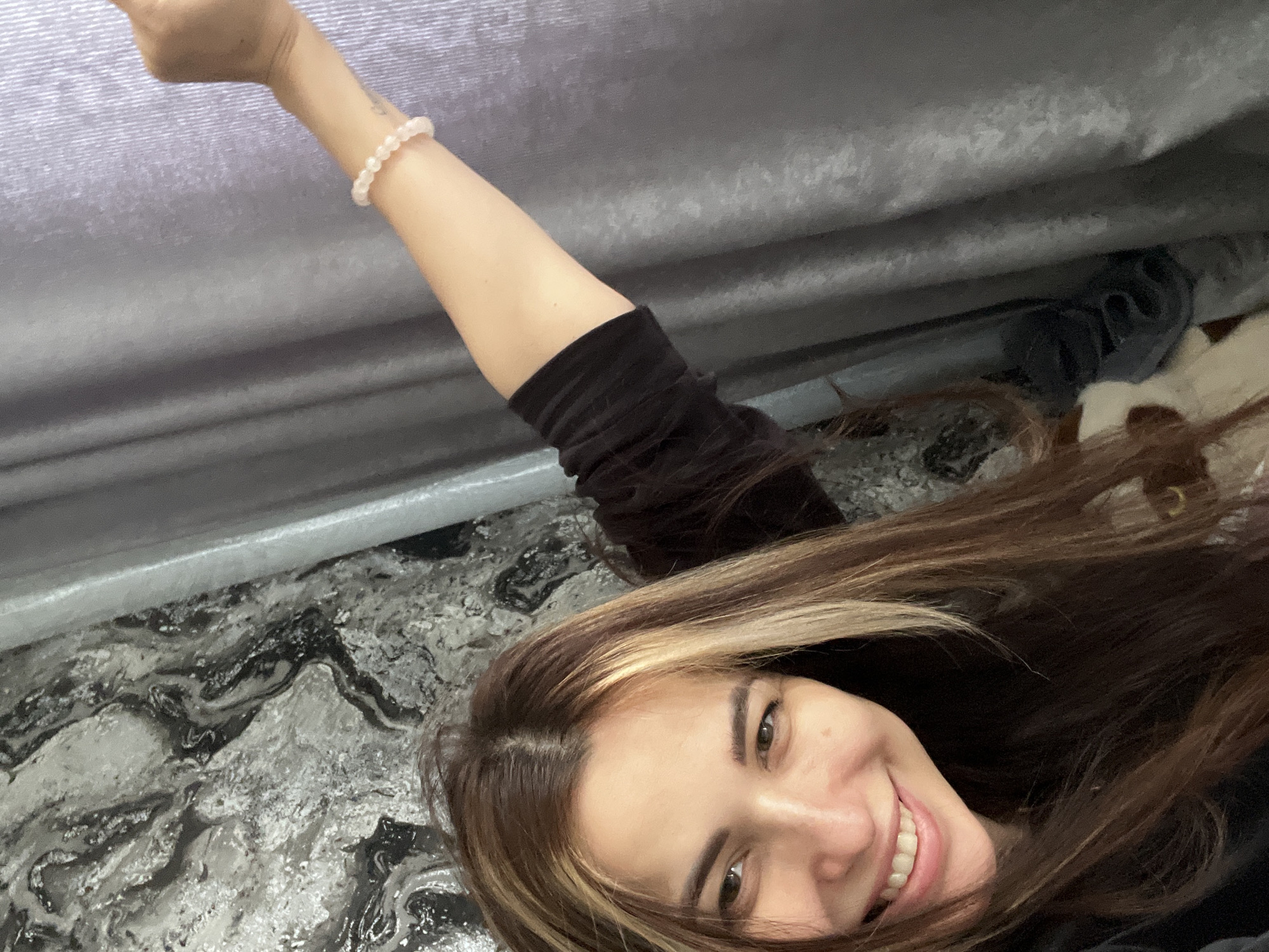 A woman with shoulder-length brown hair with blonde highlights smiles broadly at the camera. She is wearing a black long-sleeved shirt and a pink beaded bracelet on her extended arm. The background includes a textured grey surface, possibly a wall or ceiling, and a patterned surface in shades of grey, black, and white, which could be a countertop or floor. The location and city are Unknown, Unknown.