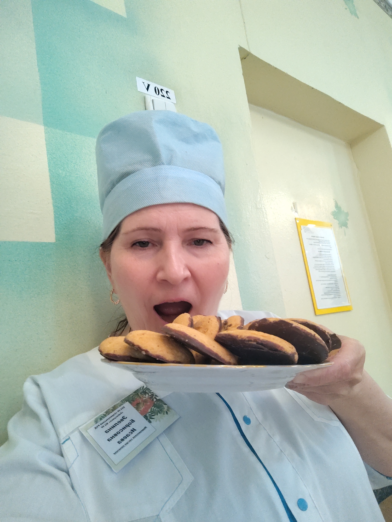 A woman, dressed in a light blue chef's hat and a white chef's coat with blue piping and buttons, holds a white plate filled with cookies. She appears to be mid-action, with her mouth wide open as if about to bite into one of the chocolate-dipped cookies. A small golden hoop earring is visible on her left ear.
The setting is indoors, likely a professional kitchen or canteen, characterized by a two-tone wall painted light green/teal and light yellow/beige. Artificial lighting illuminates the scene.
On the wall above her head, a white piece of paper is affixed, bearing upside-down text that reads "V OSS" and "350 A". To the right, a document in a yellow frame is hanging on the wall, its text illegible.
A name tag or badge is pinned to her chef's coat. The text on the badge is upside down in the image. When corrected, it reads in Cyrillic: "Для всех категорий соблюдать чистоту. Завтрак." (For all categories, observe cleanliness. Breakfast.) followed by the name "Елена Зубкова" (Elena Zubkova). The presence of the name tag and the professional attire, along with the location context of Sarapul, Russia, suggest she might be a food service worker.