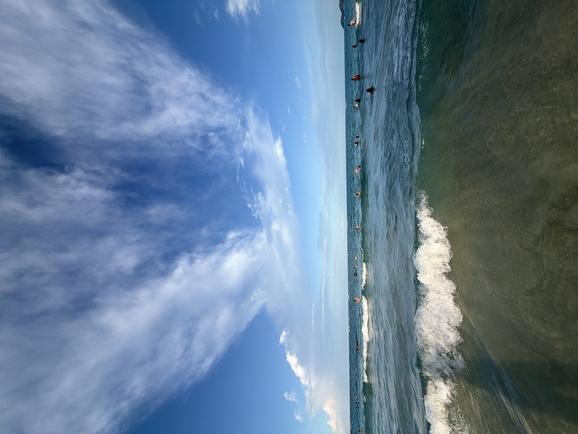 The image depicts a wide shot of a beach in Ko Samui, Thailand. The foreground is dominated by a breaking wave and the sandy seabed beneath the clear, shallow water. Beyond this, the turquoise ocean stretches towards the horizon, populated by numerous swimmers and bathers of various ages. The water is calm with gentle waves breaking closer to the shore. The sky above is a vibrant blue, adorned with wispy white clouds that are spread across the frame, suggesting fair weather. The horizon line is clearly defined, with a hint of land visible in the far distance to the right. The overall scene is one of a leisurely day at the beach.