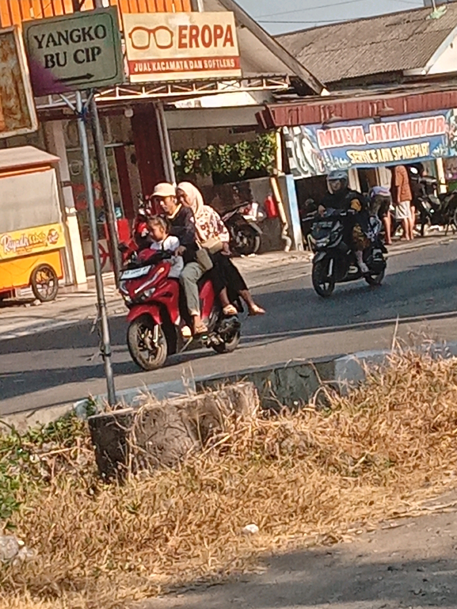 The image depicts a street scene in Banguntapan, Indonesia, likely near a commercial area. In the foreground, dry grass and a curb are visible. A grey pole stands prominently, supporting a green sign with white text that reads "YANGKO BU CIP" and an arrow pointing left. Adjacent to it is a sign with a logo of glasses and the text "EROPA JUAL KACAMATA DAN SOFTLENS".
Further into the scene, a red motorcycle is moving to the left. It carries three people: a man in a light-colored hat driving, a child seated in front of him, and a woman in a hijab and patterned shirt behind him. They are all looking forward or slightly to the side.
In the background, a building with a red tiled roof and a sign that says "MULYA JAYA MOTOR SERVICE & SPAREPART" is visible. Another person is seen on a dark-colored motorcycle, also heading towards the left. To the far left, a yellow cart with a sign partially obscured by the pole is present. The overall environment suggests a busy street with shops and ongoing traffic. The lighting indicates it is daytime.