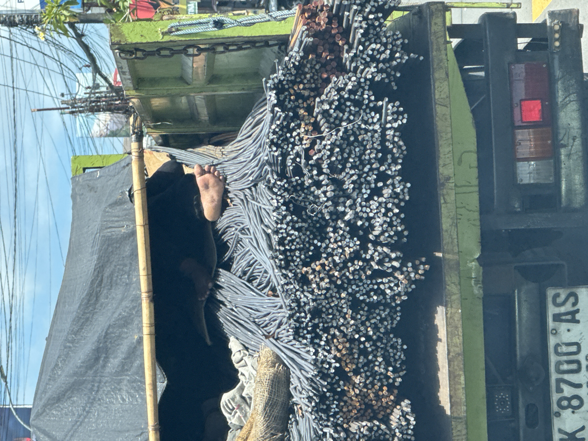 The image captures a green flatbed truck in Kuta Selatan, Indonesia, heavily loaded with steel reinforcing bars (rebar). A foot is visible, apparently belonging to a person lying among the rebar, possibly for transport. A black tarp is partially visible along the left side, seemingly covering part of the load. The truck's taillight and license plate, "K 8700 AS," are visible on the right side. Power lines are visible above, suggesting an urban or suburban setting.