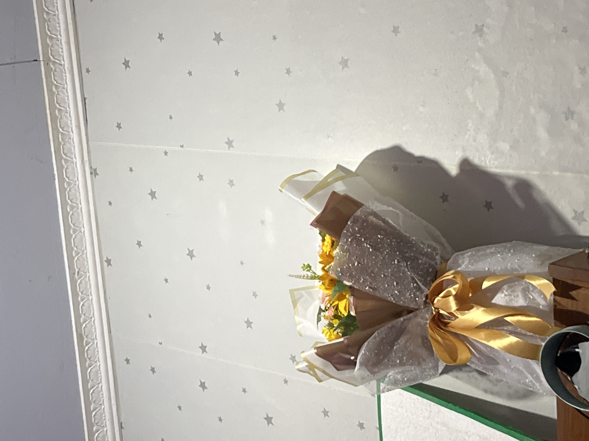 A bouquet of yellow and pink flowers, wrapped in white and brown paper with a gold ribbon, is displayed against a white wall with a subtle star pattern. The bouquet is placed on a wooden surface, with a glimpse of a green-bordered object to its left. The wall features decorative molding at the top. The lighting suggests an indoor environment, possibly a home, with a shadow cast on the wall from the bouquet and an unseen object. The image is taken from a low angle, looking upwards towards the wall and ceiling. The location is identified as Glagah Kulon, Indonesia.