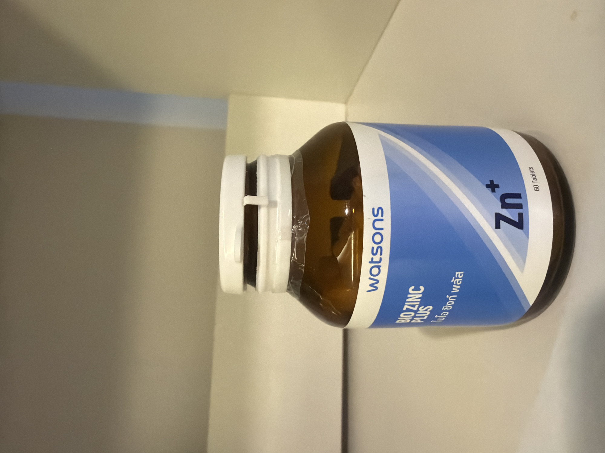 A brown plastic bottle of Watsons Bio Zinc Plus supplements sits on a white shelf. The bottle has a white cap and a blue label with white text. The label reads "watsons" at the top, followed by "BIO ZINC PLUS" and Thai text. At the bottom of the label, "Zn+" is printed in large blue letters, with "60 Tablets" in smaller white text below it. The background is a blurred off-white shelving unit. The image appears to be taken indoors, in what could be a pharmacy or a home storage area. The location is not specified in the image.