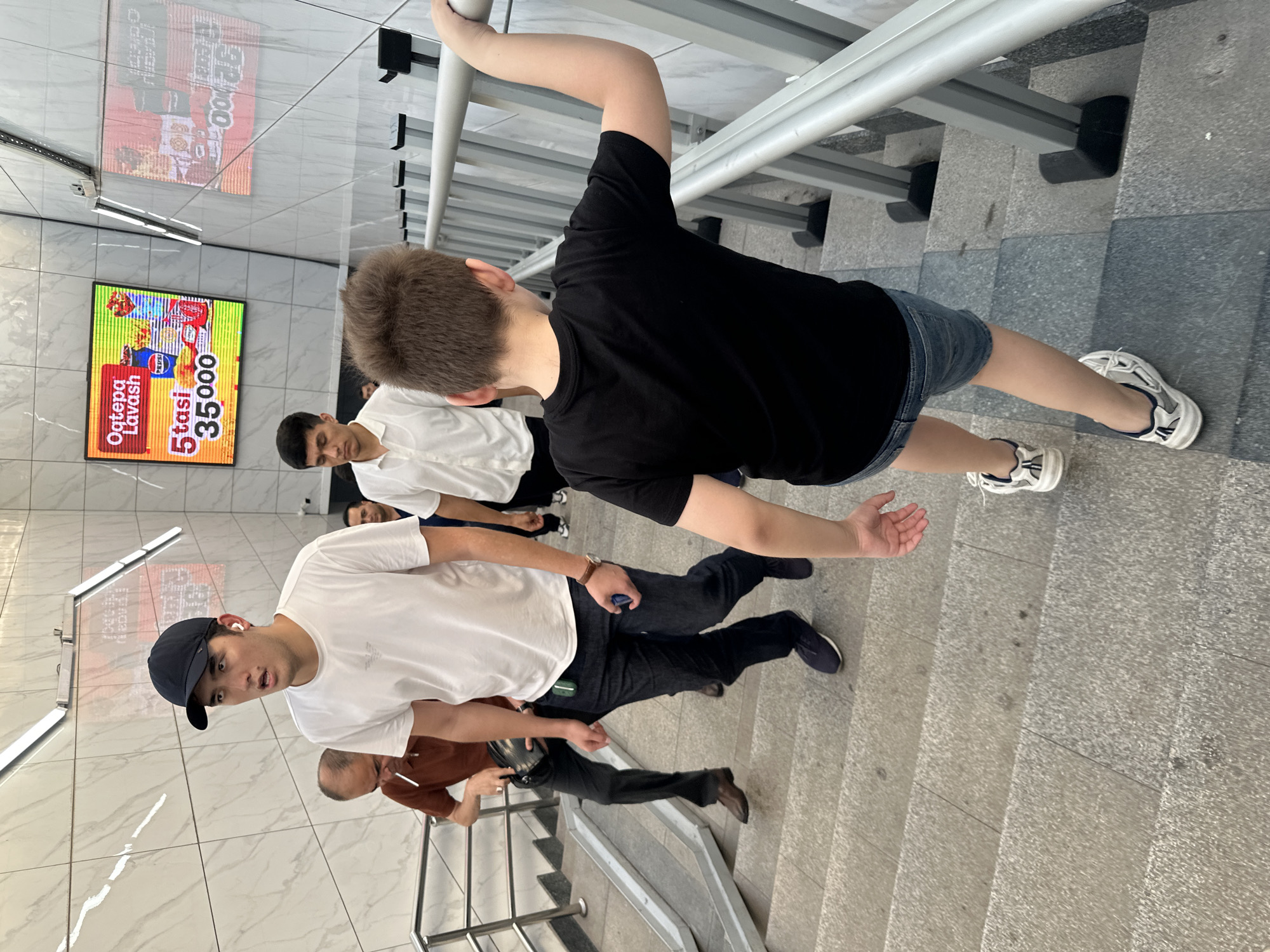 The image depicts a group of individuals ascending a wide, modern concrete staircase in Tashkent, Uzbekistan. The architecture features polished marble-tiled walls and a metal railing system. Dominating the background is a large digital advertisement displaying food promotions, including images of fast food items and text in Uzbek.
In the foreground, a young person with light brown hair is seen from behind, wearing a black t-shirt and denim shorts, holding onto the railing. To their left and further up the stairs, a young man in a white t-shirt and dark jeans, wearing a black baseball cap and white earbuds, looks to his left with an open mouth, as if surprised or speaking. To the left of him, an older man with balding grey hair is walking up the stairs, holding a cigarette in his right hand and a dark bag in his left. Further up the stairs and behind the young man in the white t-shirt, another young man in a white collared shirt is visible. Several other individuals are partially visible on the stairs. The lighting suggests an indoor environment, likely a public transit station or a commercial building.