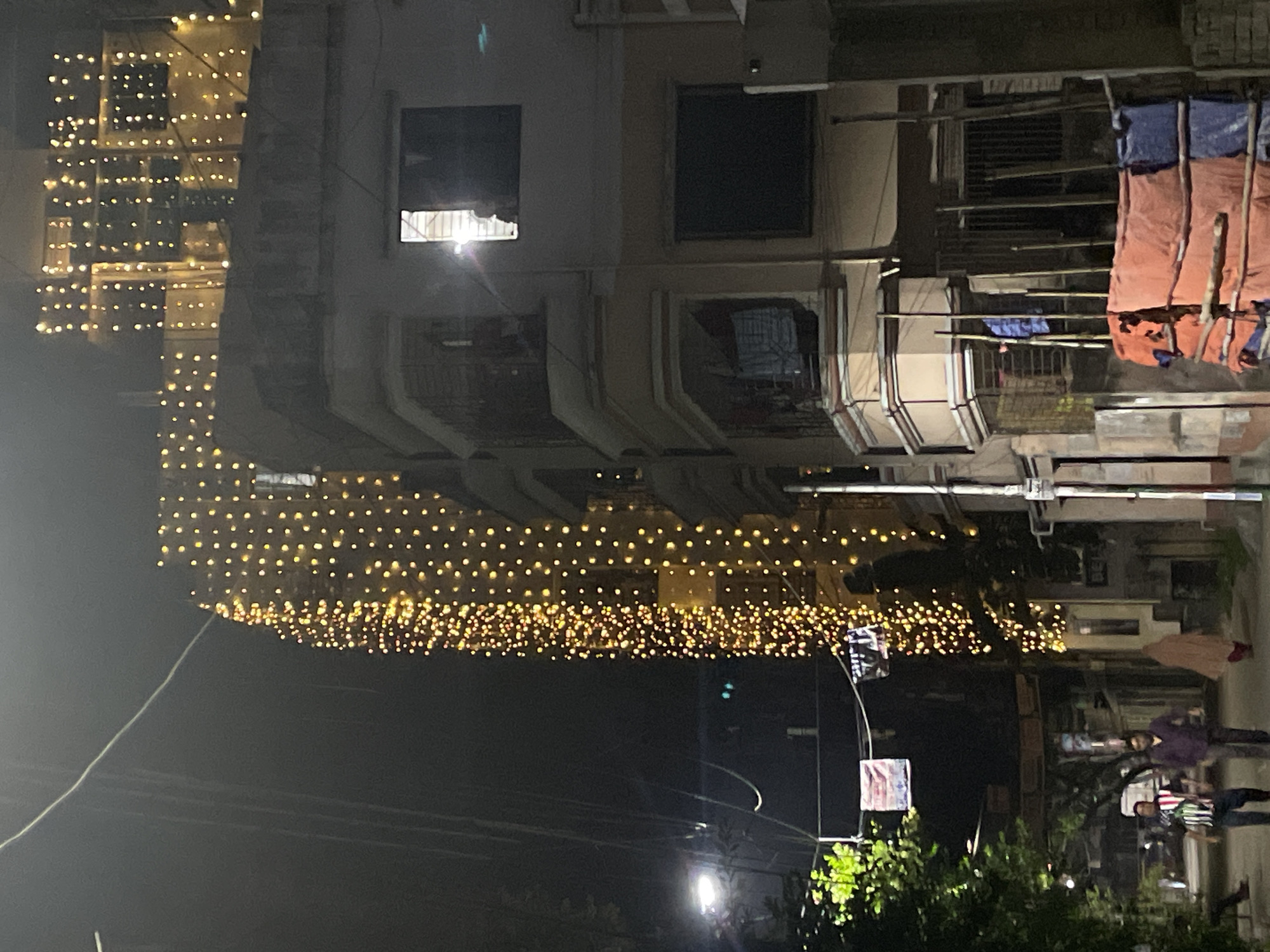 The image depicts a street scene at night, likely in Dhaka, Bangladesh, based on architectural cues and cultural context. Buildings, possibly apartment blocks, are visible on both sides of the street. The facades are adorned with strings of small, warm-toned lights, suggesting a festive or celebratory occasion. Some laundry hangs from balconies on the right. Below, streetlights illuminate a few people walking, and foliage from trees is visible. Signboards are positioned near the right, partially obscured by tree branches. The overall scene conveys a sense of nighttime activity and festive decoration.