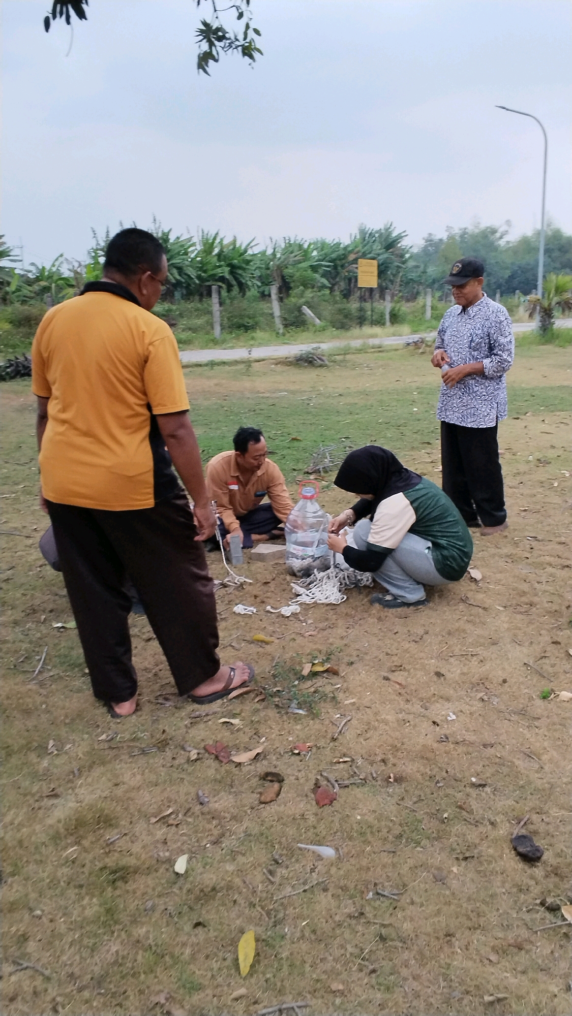 A group of four individuals is gathered outdoors in what appears to be a park or open field. The ground is covered in dry grass and scattered leaves. In the background, there are rows of banana trees and a road. A street light is visible on the right side of the image.
On the left, a man wearing a yellow t-shirt and dark pants stands facing away from the camera. To his right, another man in an orange shirt and dark pants is seated on the ground. In front of him is a large clear plastic water bottle and a pile of white rope. A woman wearing a hijab and a green and beige shirt is crouched down, actively working with the rope. To the far right, a man in a patterned shirt and a dark baseball cap stands holding a small object, possibly a bottle. The overall atmosphere suggests a casual outdoor activity. The location is identified as Mojokerto, Indonesia.