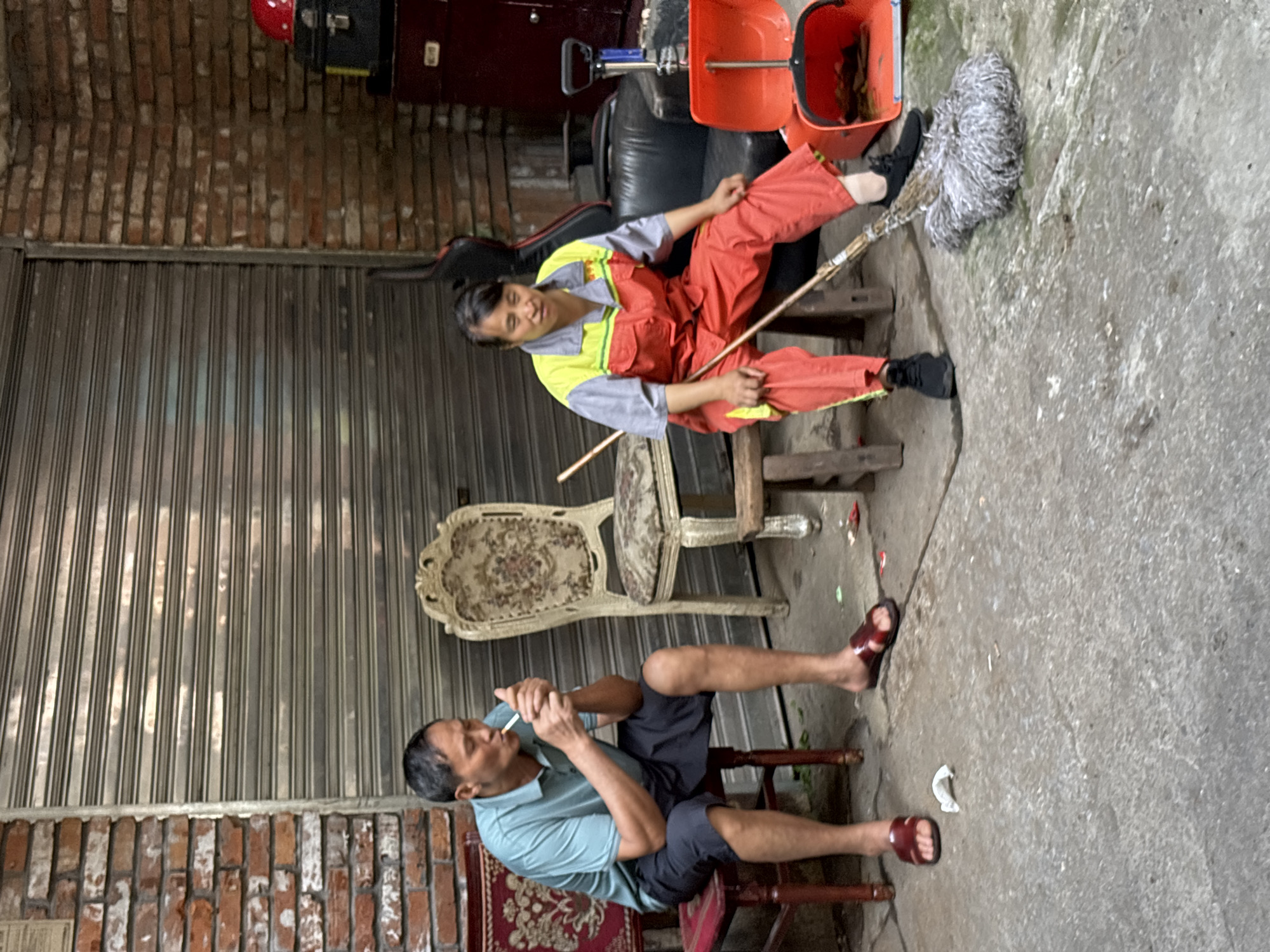 The image depicts two individuals in what appears to be an outdoor, urban setting, possibly a back alley or workspace. In the foreground, a man is seated on a red chair, smoking a cigarette. He is wearing a light blue polo shirt and dark shorts. His expression is neutral, and he is focused on lighting his cigarette.
Behind him, slightly to the right, a person in an orange and grey uniform, likely a cleaner or worker, is seated on a wooden stool. They are holding a mop with a gray head and a wooden handle, and appear to be resting or taking a break. The uniform is a bright orange and grey combination, with a yellow accent on the collar. The person is looking downwards and slightly to the side.
The scene is characterized by a weathered metal roll-up door in the background, and a brick wall to the left. An ornate, possibly antique, chair with patterned upholstery is visible between the two seated individuals. To the upper right, an orange bucket and part of a red object, possibly a helmet or tool, are visible. The ground is concrete, with some debris scattered around.
The overall impression is one of a pause in daily work, with one person taking a moment to smoke and the other seemingly resting with their cleaning equipment nearby. The lighting suggests it is daytime, though the exact time is not discernible. The location cues like the brickwork and the style of the metal door, along with the provided context of Yuzhong District, China, suggest an urban environment in that region. No visible text is present in the image.