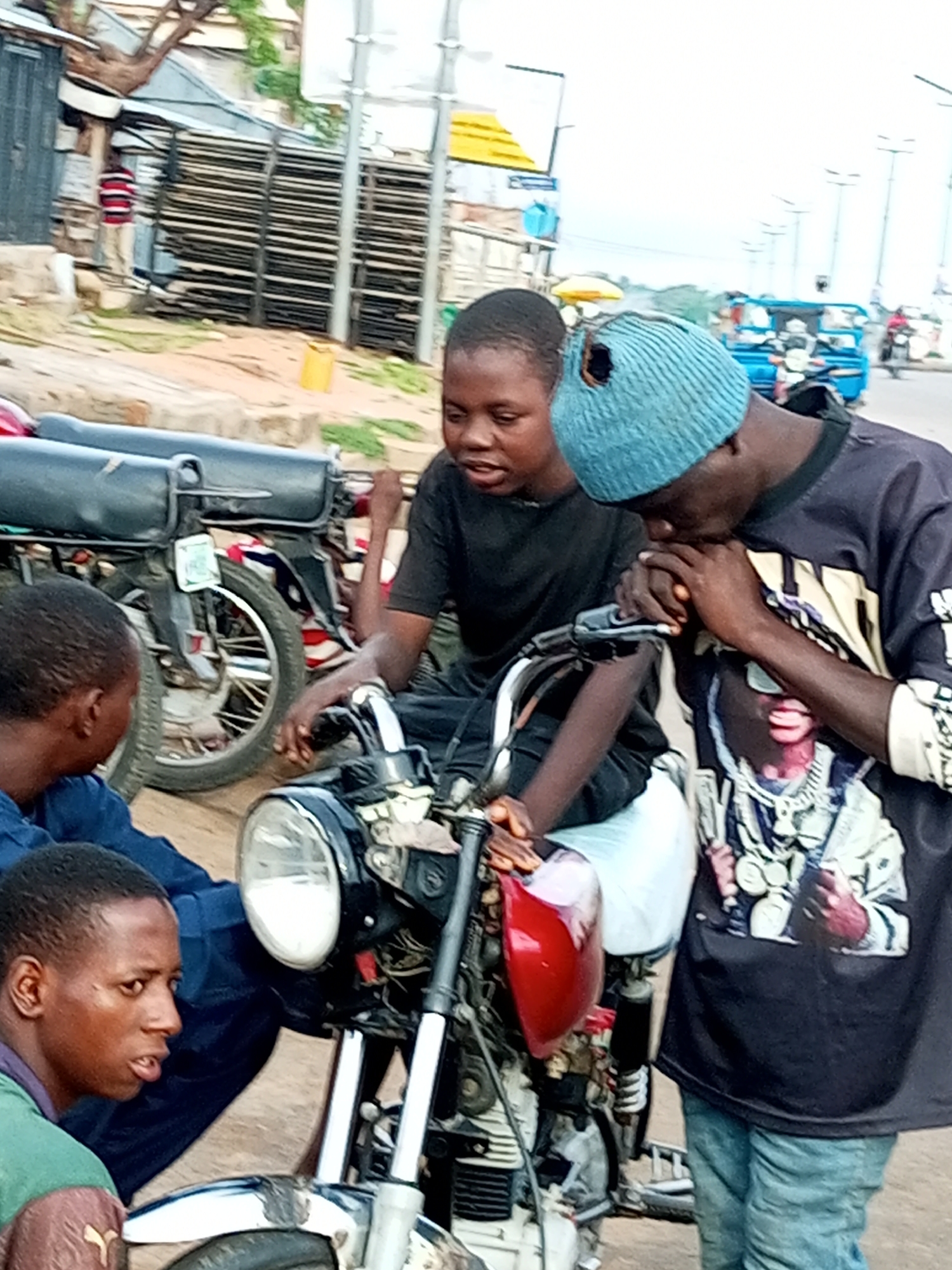 In Gombe, Nigeria, three young males are gathered around two motorcycles on a dirt road. The central figure, wearing a black t-shirt and a light blue beanie with a hole in it, leans forward, resting his hands near the handlebars of the motorcycle in the foreground. To his left, another young male in a black t-shirt looks towards the foreground, his mouth slightly open. A third individual, seen from the left side of the image, is partially visible, wearing a blue jacket and looking downwards. The motorcycles appear to be the main focus, with parts of their engines, wheels, and seats visible. In the background, a building with wooden planks is situated, and a yellow plastic container is on the ground. The overall environment suggests a roadside setting in a semi-urban area.