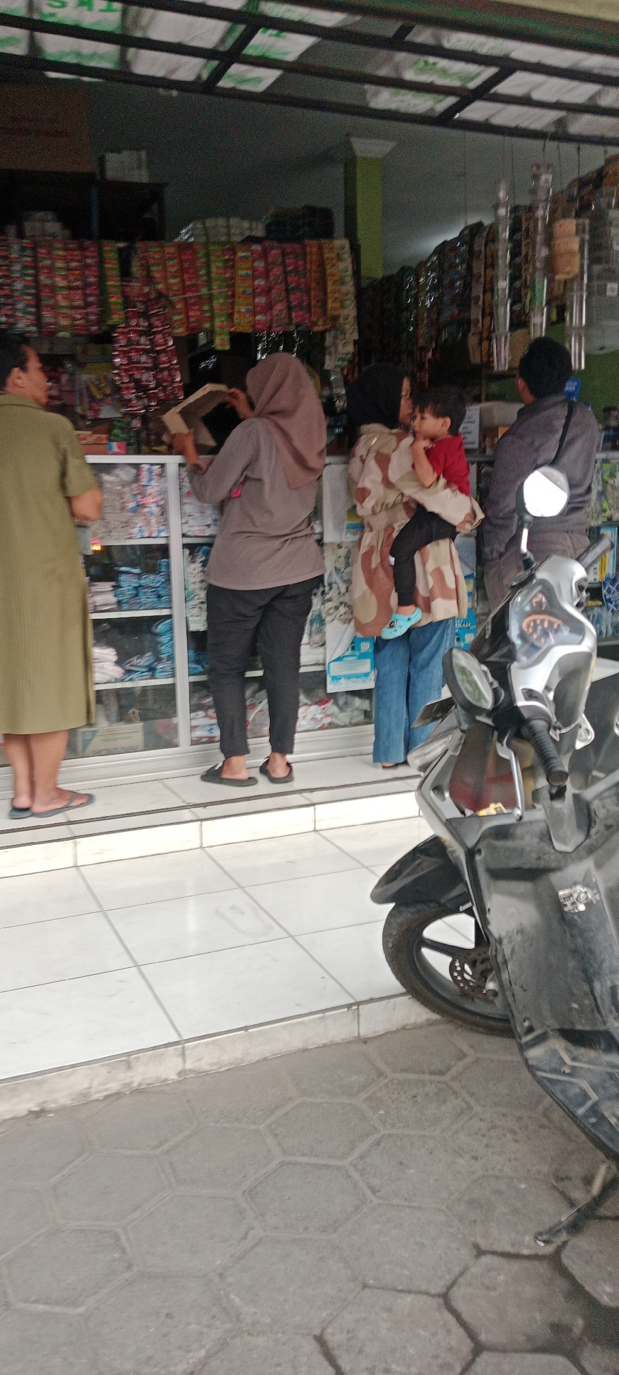 In Maguwoharjo, Indonesia, a street-level view captures an outdoor scene in front of a small retail establishment. Four individuals are visible, with a motorcycle parked in the foreground to the right.
On the left, a person wearing a long, olive-green dress stands with their side profile visible. To their right, a person in a taupe hijab and dark pants stands behind a glass display case. This individual is holding a light brown cardboard box and appears to be examining its contents. Further to the right, a person in a patterned long-sleeved top and blue jeans holds a young child. The child, wearing a red shirt and light blue footwear, is facing left. Behind them, a person in a dark grey jacket stands with their back to the viewer.
The retail establishment features shelves stocked with various packaged goods. The pavement in front of the store is tiled with white square tiles, transitioning to hexagonal paving stones in the foreground. A dark-colored motorcycle is parked on the paving stones, with its front wheel and part of the body visible. The overall lighting suggests it is daytime.