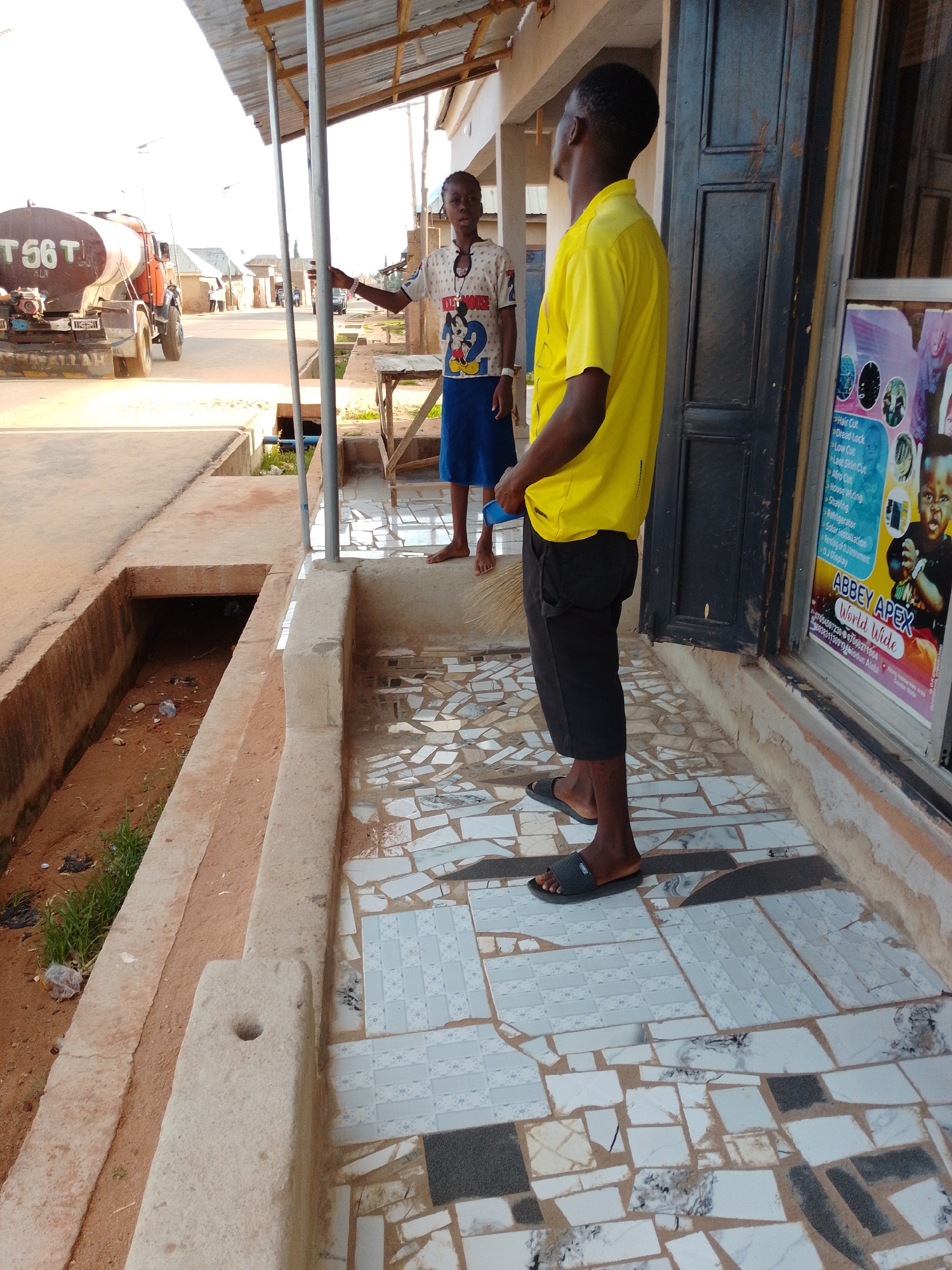 The image depicts a street scene in Gombe, Nigeria. Two individuals are present. In the foreground, a young man wearing a yellow t-shirt and black shorts stands facing away from the camera, looking towards a building with a dark-colored door. He is wearing black flip-flops and holding a broom in his right hand. To his left, a younger person, possibly a teenager, is standing in front of the same building, wearing a white t-shirt with a Mickey Mouse design and a blue skirt. They are looking towards the street.
A truck with a large tank, marked "T 56 T", is visible in the background on the left side of the image, driving on the street. The ground in the foreground is paved with mosaic tiles in various shades of white, grey, and black. To the left of the paved area, there is a drainage ditch filled with sand and some debris. The building has a corrugated metal roof supported by metal poles. A sign is visible on the window of the building, featuring text and images related to hair services, including "ABBEY APEX World Wide". The overall lighting suggests it is daytime, with sunlight casting shadows.