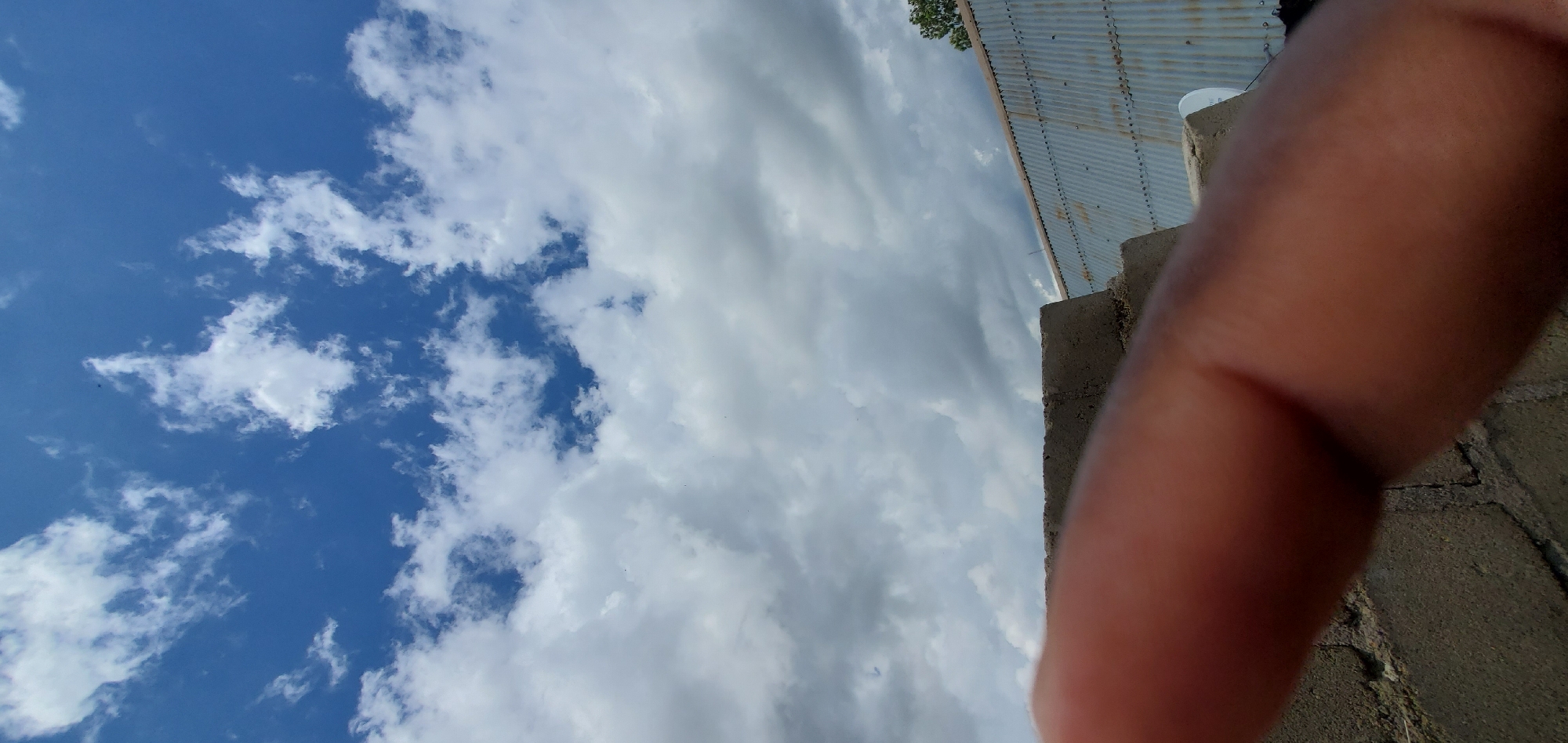 A close-up of a human finger is in the foreground, partially obscuring a wall made of concrete blocks. Beyond the wall, a corrugated metal roof is visible, and a single tree branch with green leaves is seen on the right. The background is dominated by a sky with scattered white and gray clouds. The overall scene appears to be outdoors, possibly on a rooftop or elevated surface, given the presence of building materials and the sky.