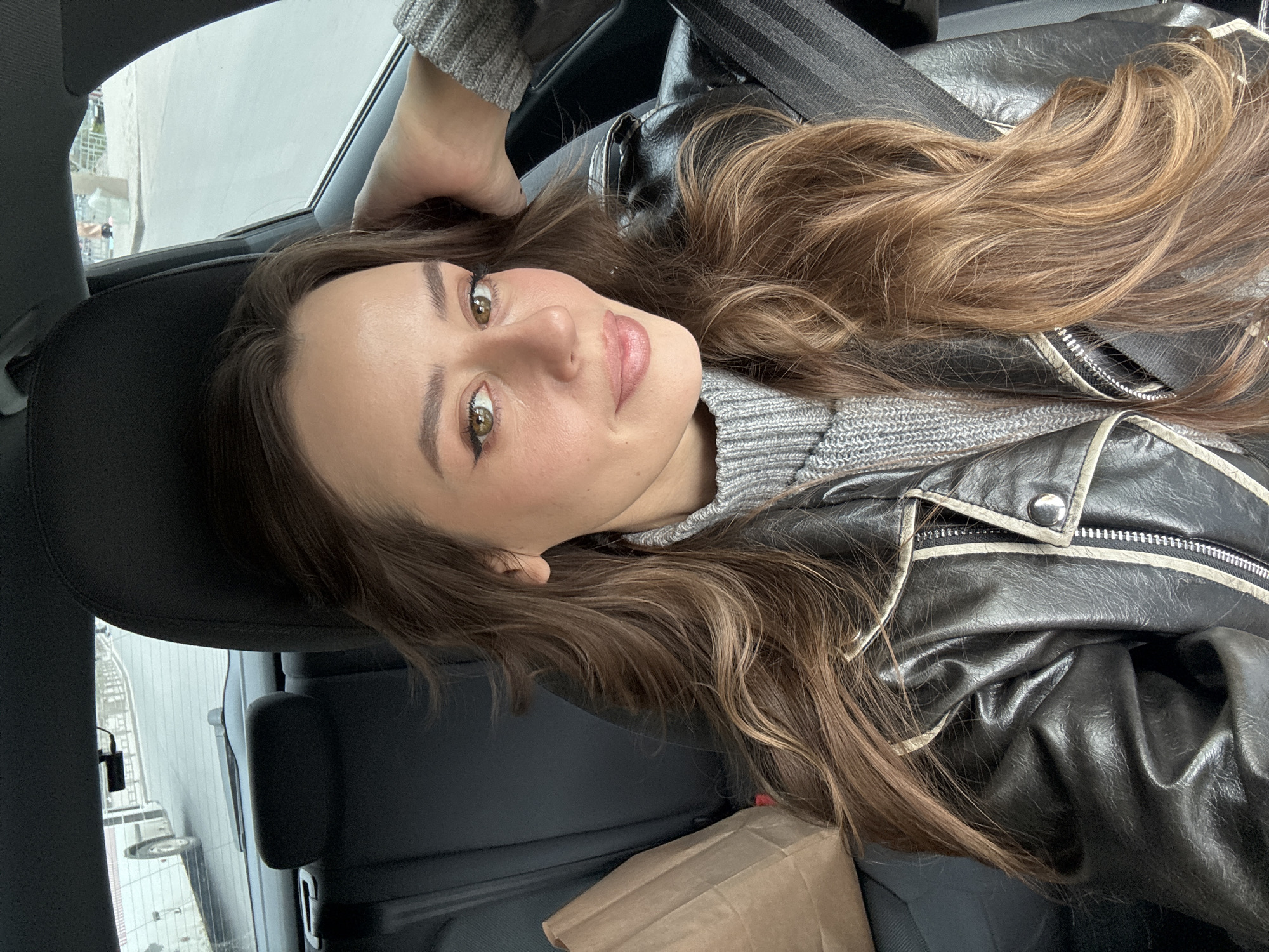 A young woman is reclined in the driver's seat of a car, taking a selfie. She has long, light brown hair and is wearing a grey sweater and a black leather jacket. The car is parked on a street in Krasnoyarsk, Russia, as evidenced by the buildings visible through the car window.