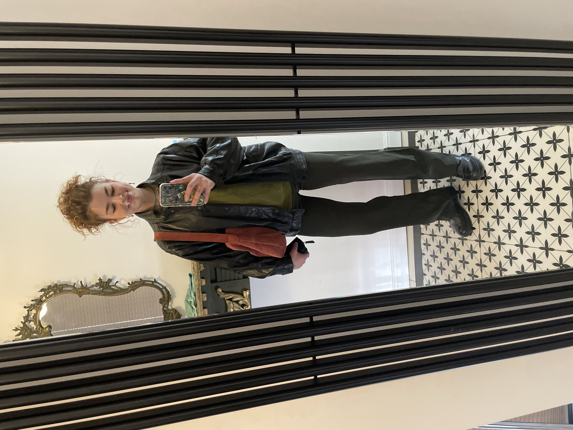 A person stands in front of a mirror, holding a phone in their right hand, presumably taking a photo of themselves. The person is wearing a black leather jacket over a green top, dark pants, and black shoes. They have curly reddish-brown hair. They are carrying an orange shoulder bag. The mirror is framed with black and white horizontal stripes. Behind the person, in the reflection, is an ornate gold-colored mirror. To the right of the person in the reflection is a patterned black and white tile floor. The location is likely indoors. There is no concrete information that indicates this is in the city of Tbilisi, Georgia.
