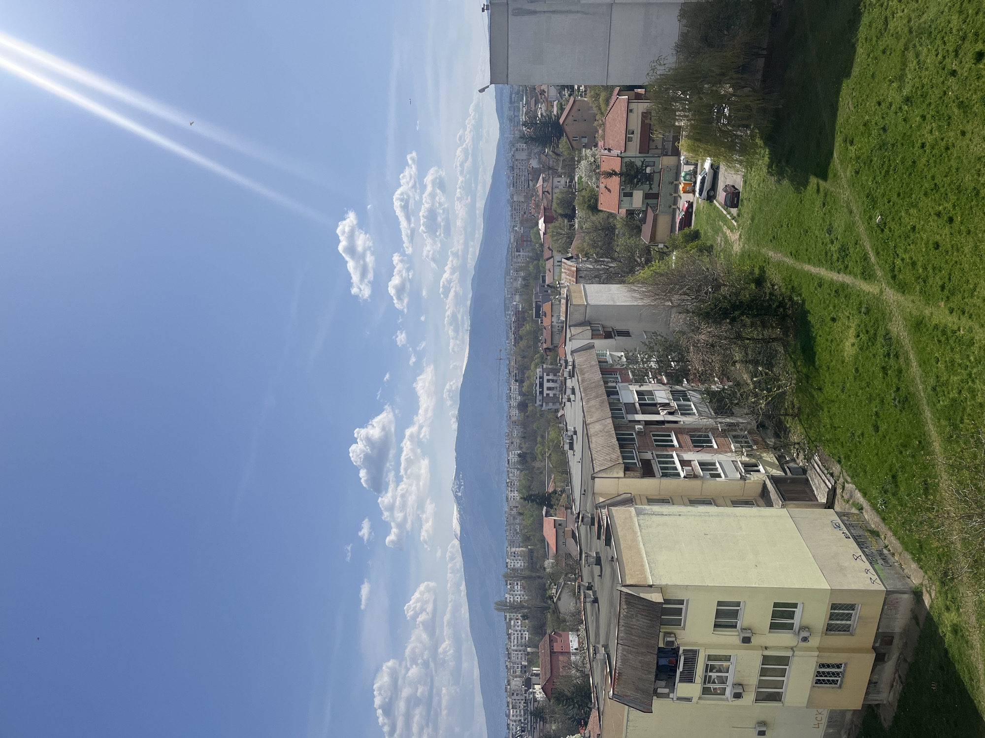 The image captures a high-angle, wide view of a residential area in Sofia, Bulgaria, under a bright blue sky.
In the immediate foreground, a vibrant green grassy area stretches across the right side, traversed by several worn dirt paths. On the left, a multi-story, pale yellow apartment building stands prominently, featuring numerous windows and external air conditioning units. Faint dark graffiti is visible on its side wall, with partially legible Cyrillic text including "БЕЗ *** ЦЕНТРАЛНА ПЛАНИРАЩА" (WITHOUT *** CENTRAL PLANNING) and the letters "ЧСК" at the lower right.
The midground reveals a dense urban landscape comprising various residential buildings, including apartment blocks and houses with red-tiled roofs, interspersed with trees and small green spaces. Several cars, including white, red, and dark-colored vehicles, are parked near some of the houses.
In the distant background, a long mountain range, characteristic of Vitosha Mountain near Sofia, dominates the horizon, with subtle hints of snow on its peaks. The sky above is a clear, brilliant blue, adorned with scattered white cumulus clouds. The sun is visible in the upper left quadrant, emitting distinct, bright rays. A small, dark, winged object (possibly a bird or small aircraft) is visible high in the sky, along with another tiny speck.
The scene depicts a sunny day, likely morning or early afternoon, indicated by the strong sunlight and visible shadows. The overall setting is a bustling urban environment framed by a prominent natural landscape.