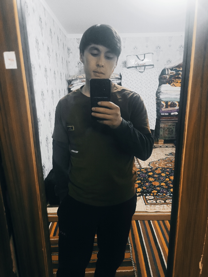 A young man stands in front of a mirror, taking a selfie with his smartphone. He is wearing a dark green t-shirt over a black long-sleeved shirt and black pants. The background includes patterned wallpaper, a neatly stacked pile of bedding or clothes, and a colorful rug with floral designs. The mirror's wooden frame is visible on both sides, and the floor is covered with a striped rug.