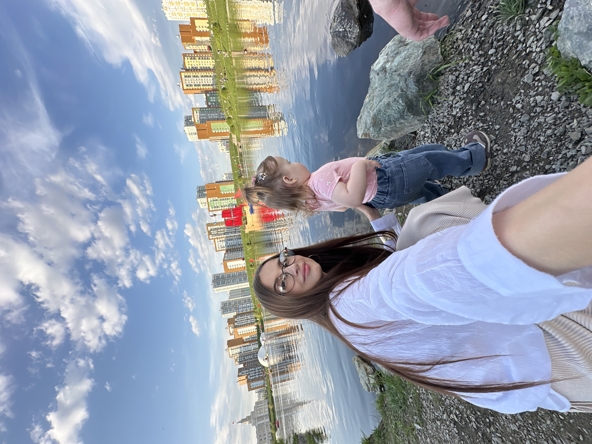 A selfie captured by a woman with long brown hair and glasses, wearing a white shirt, features a young girl standing next to her. The girl is dressed in a pink shirt and jeans. They are situated near a body of water, likely a river or lake, with pebbles and rocks scattered around. In the background, a clear blue sky with fluffy clouds is visible, along with the reflection of city buildings in the water. The buildings suggest that the location is in the city of Yekaterinburg, Russia.
