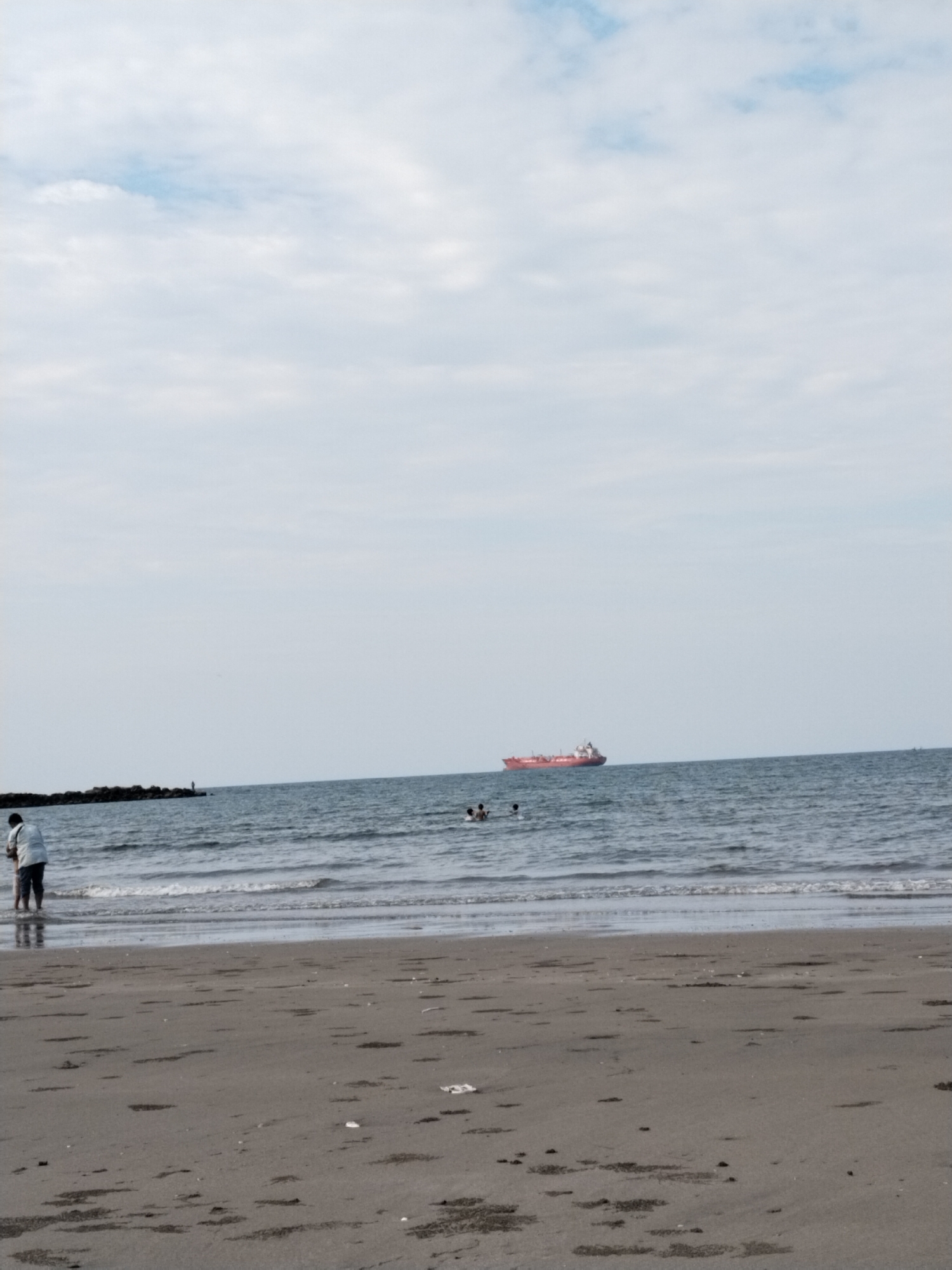 This image depicts a tranquil beach scene in Lhokseumawe, Indonesia, under an overcast sky with light, hazy clouds. The foreground features a dark sandy beach, showing patches of dampness and scattered debris.
In the lower-left, a person wearing a light-colored shirt and dark pants stands in the shallow water near the shore, possibly with a child whose outline is partially obscured by the adult's body. The calm ocean extends into the mid-ground, with gentle waves breaking near the shoreline. Further out in the water, towards the center-right, two individuals appear to be swimming or wading.
On the left side of the mid-ground, a dark rocky breakwater or jetty stretches into the sea, with a small, indistinct figure visible at its far end, likely a person fishing. A prominent feature in the mid-distance is a large red cargo or tanker ship, sailing from left to right across the calm blue waters.
The overall scene suggests a leisurely daytime activity at the beach. No specific text is visible in the image.