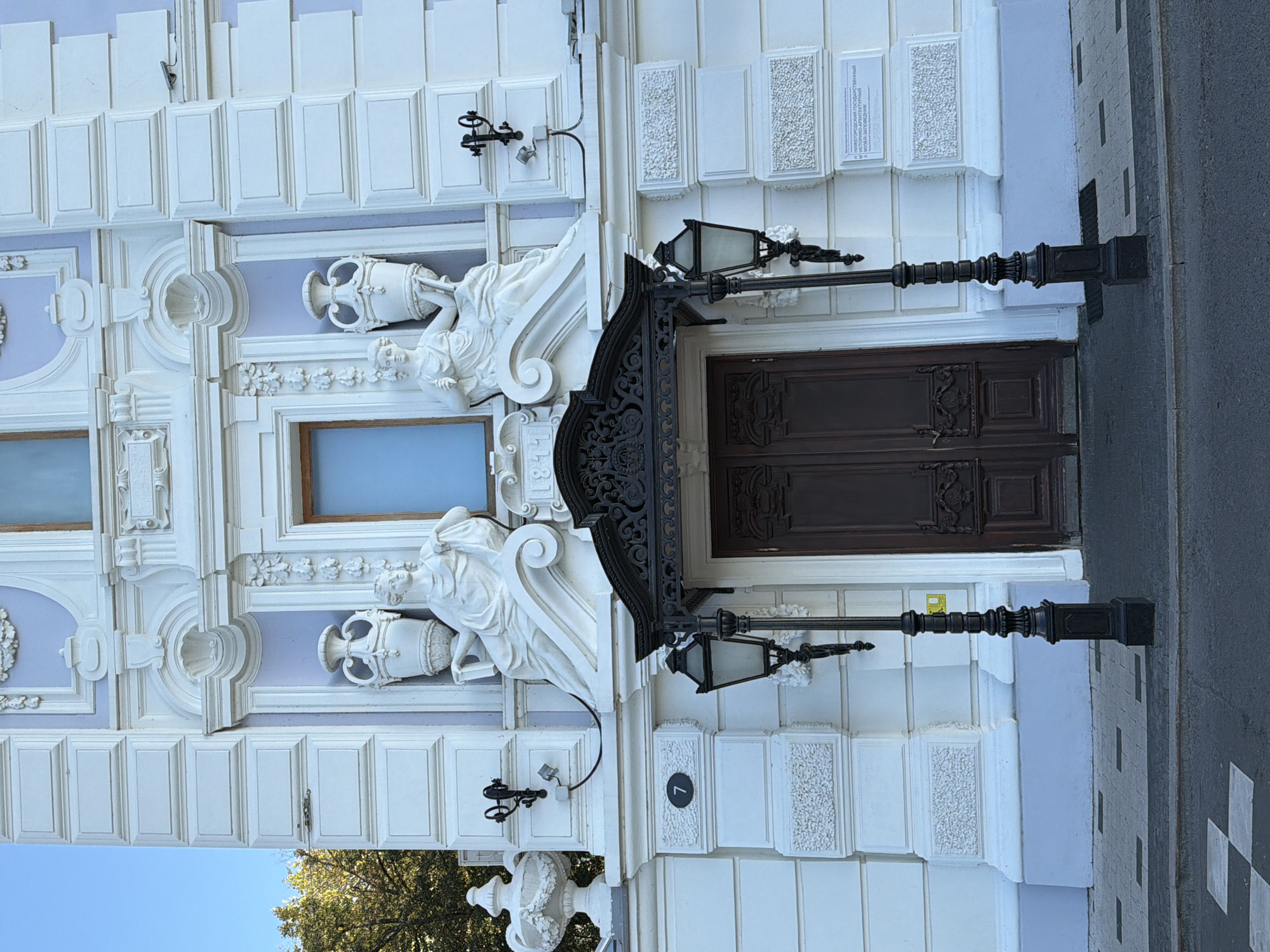 The image shows the entrance of the Nizhny Novgorod State Architectural and Historical Museum-Reserve, located at house number 7. The facade of the building is ornate and white, with light blue accents. Two neoclassical statues of women holding urns flank a large, dark wooden double door. Above the door, a wrought-iron canopy is visible, and two vintage-style lampposts stand on either side of the entrance. The building is situated on a street with a checkered pavement and a clear blue sky above.