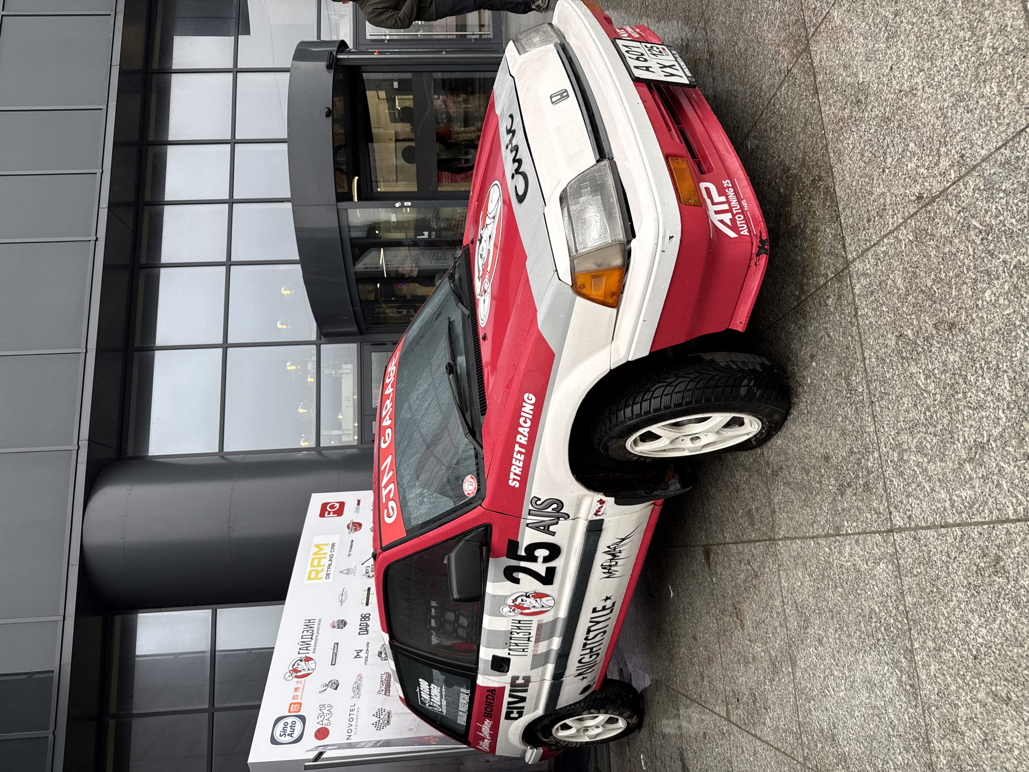 The image depicts a red and white Honda Civic race car parked on a wet sidewalk in front of a modern building with glass windows. The car is adorned with racing decals, sponsor logos, and the number "25." A sign to the left of the car displays the logos of various companies, including "Sino Auto," "NOVOTEL VLADIVOSTOK," "RAM DETAILING CAR," and "GIN GARAGE." A person is partially visible at the entrance of the building, suggesting a potential event or gathering. Based on the signage, the location is Vladivostok, Russia.