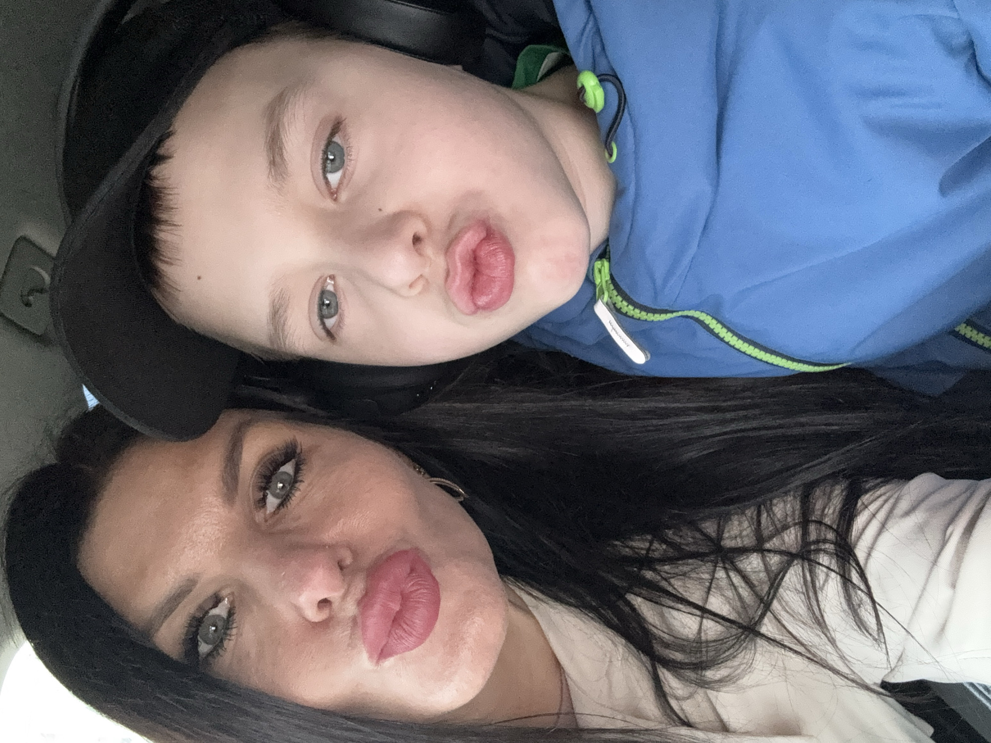 A selfie-style photograph depicts two individuals, a woman and a child, likely a boy, posing closely together. Both are exhibiting a "pursed lips" expression. The woman is in the lower portion of the frame, her head tilted, with long, dark hair. She has light skin and appears to be wearing makeup. A light-colored garment is visible. The child is positioned above her, wearing a blue jacket with green detailing and a black baseball cap. Both have light-colored eyes. The specific location in the city of Chaikovsky, Russia, cannot be determined from this image.
