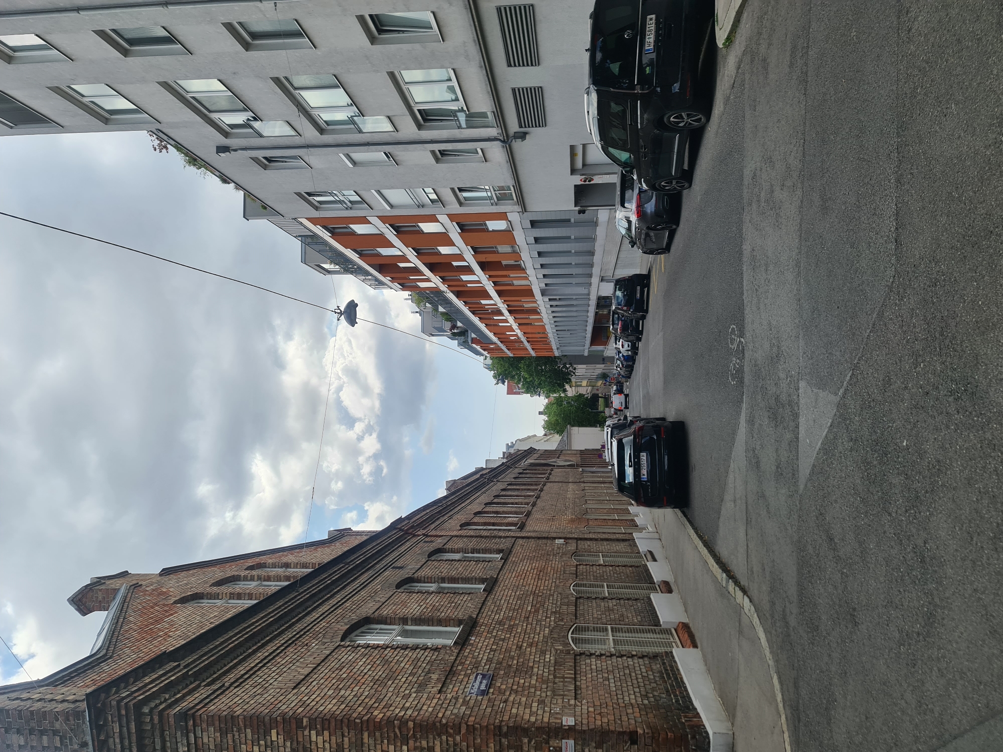 The image depicts a street scene in Vienna, Austria, characterized by a mix of old and new architecture. On the left, a weathered brick building with arched windows lines the street. To the right, modern apartment buildings with gray and orange facades rise higher. Several cars are parked along the street, including a black SUV in the foreground with license plate "HF 581EW" and a black station wagon with license plate "W 25517 H". The sky is overcast with scattered clouds. The street is paved, and a sidewalk is visible along the left side of the road, marked with a street sign reading "Michelbeuern-Gasse". A bicycle lane marking is present on the road. The perspective is from ground level, looking down the street.