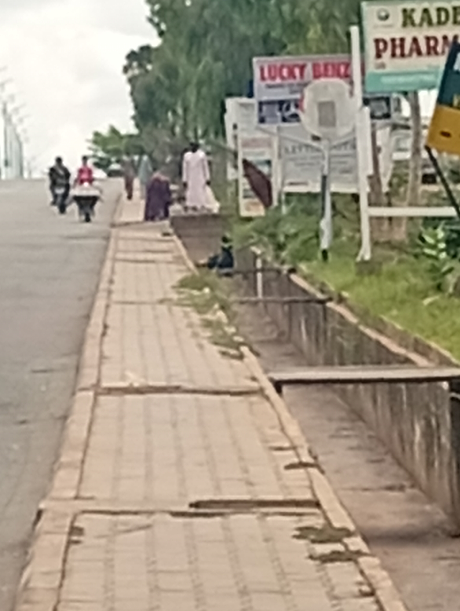 The image captures a street scene in Gombe, Nigeria, with a focus on a sidewalk adjacent to a road. A concrete gutter runs along the side of the sidewalk, bordered by overgrown grass. On the road, several individuals are visible. Two people are riding motorcycles in the distance, and further down the road, a person in a white robe stands near a purple-robed figure and another person in a white robe. In the background, various signs are posted, including one for "KADE PHARM" and another for "LUCKY BENZ." A person is seated on the edge of the gutter, facing away from the camera. The sky is overcast, suggesting a cloudy day. The sidewalk is paved with interlocking concrete blocks.