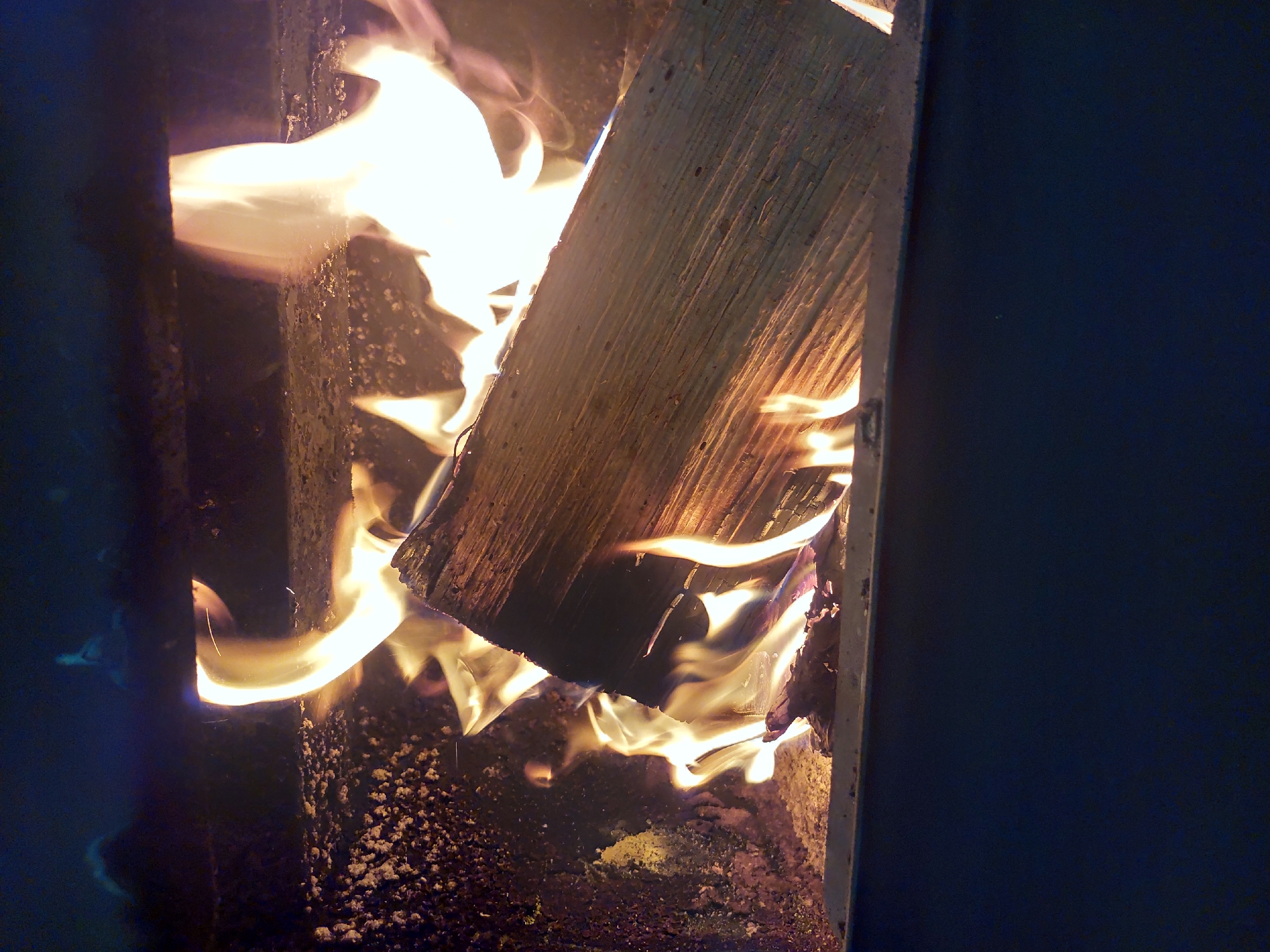 A close-up shows flames consuming wood within a wood-burning stove. The fire's bright orange and yellow light illuminates a piece of split wood held within the confines of the stove. The stove's interior surfaces appear dark and sooty, with traces of ash. The surroundings are dimly lit, with a dark blue-black backdrop. The scene is not identified with a location.