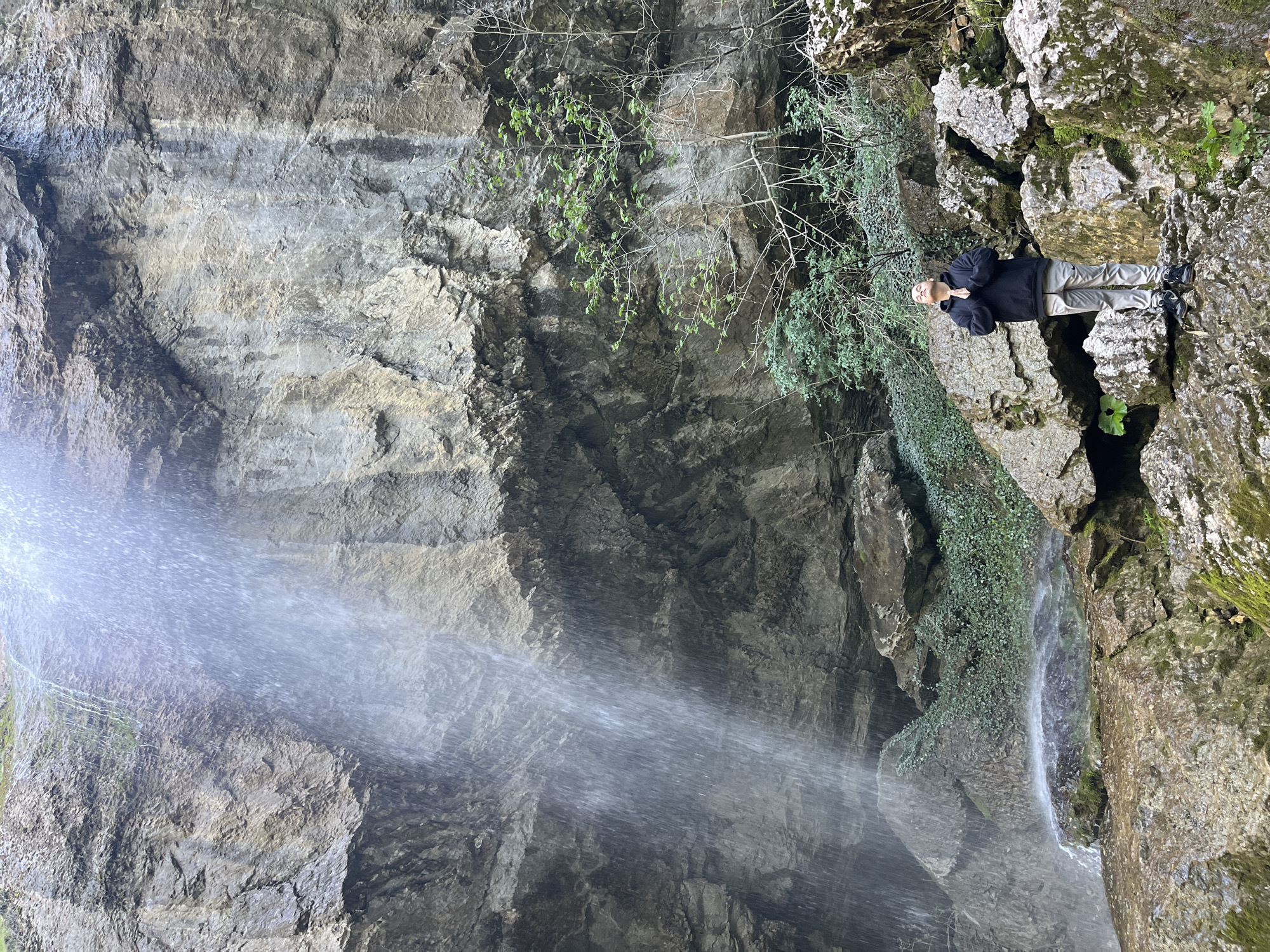 A long shot captures the interior of a large cave or rock formation, likely in Veselinovo, Bulgaria. A man in a black jacket and light-colored pants is standing on a rocky ledge, situated to the right of the frame. A waterfall cascades from above, visible in the lower left, creating a mist. Moss and vines cling to the cave walls.