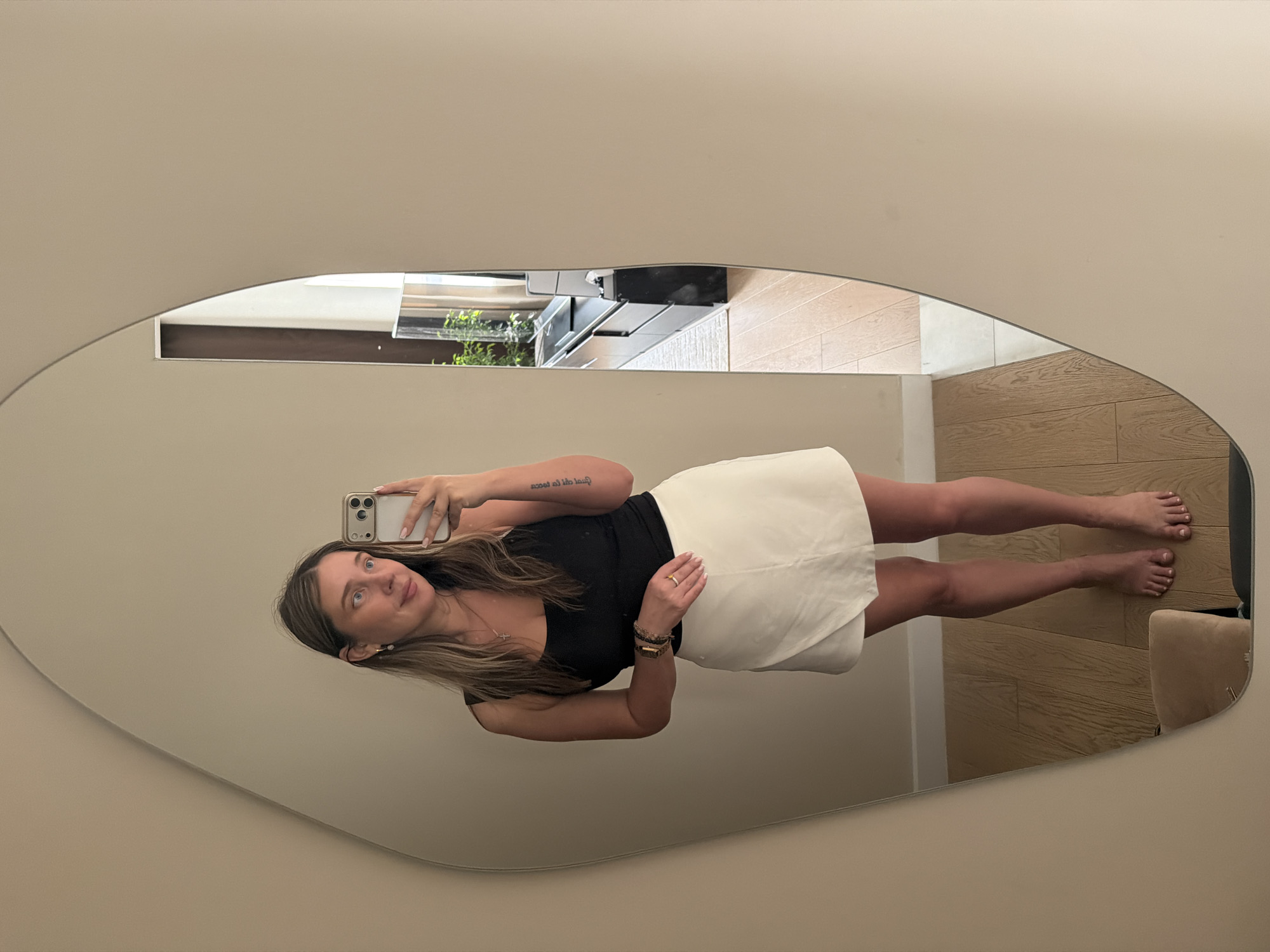 A woman stands in front of a large, irregularly shaped mirror, taking a selfie with her phone. She is wearing a black tank top and a white wrap skirt. She has blonde hair and is wearing earrings and bracelets. In the reflection of the mirror, a living room is visible, with a television, a black media console, and a large window. The floor is made of light-colored wood. The scene is indoors, likely in a residential setting. The specific location in Укромновское сельское поселение, Ukraine cannot be determined from the visual information provided.