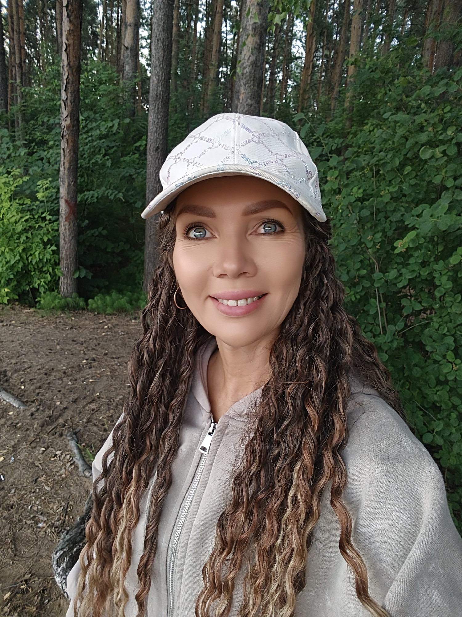 The media file is a close-up, selfie-style portrait of a woman in an outdoor setting. She is wearing a light-colored baseball cap with a subtle patterned design and a light gray zippered hoodie. Her long, curly, ombre-colored hair cascades down her shoulders and chest. She has blue eyes and is smiling at the camera, with a cheerful and relaxed expression.
The background consists of a forest with tall pine trees and dense green foliage. The ground in the foreground appears to be a mixture of dirt and fallen leaves, with some scattered branches. The lighting suggests it is daytime, likely mid-morning or afternoon, and the weather seems pleasant. The overall impression is of a casual outing in nature, possibly a walk in the woods. There is no visible text in the image.