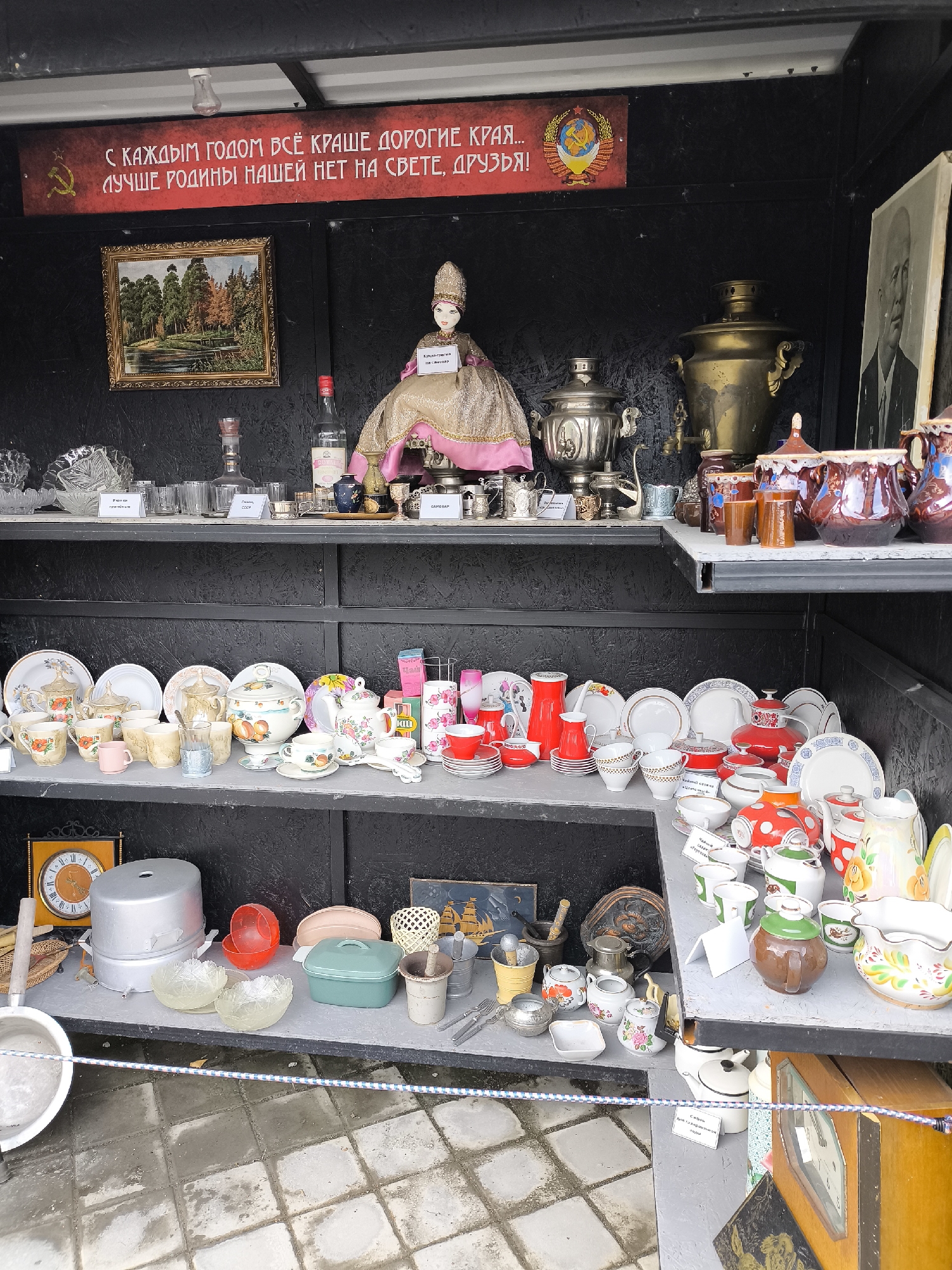 The image shows a stall at a flea market in Borgustanskaya, Russia. The stall is filled with various items, mostly vintage Soviet-era kitchenware and decorative objects. A prominent red banner with Cyrillic text and a Soviet emblem hangs above the stall. Displayed on shelves are ceramic bowls, plates, cups, teapots, and samovars. A framed landscape painting and a portrait of a man are also visible. On the lower shelf, there are more dishes, a large metal pot, a clock, and other miscellaneous items. The ground in front of the stall is paved with grey and white tiles. There are no people or animals visible in the image.