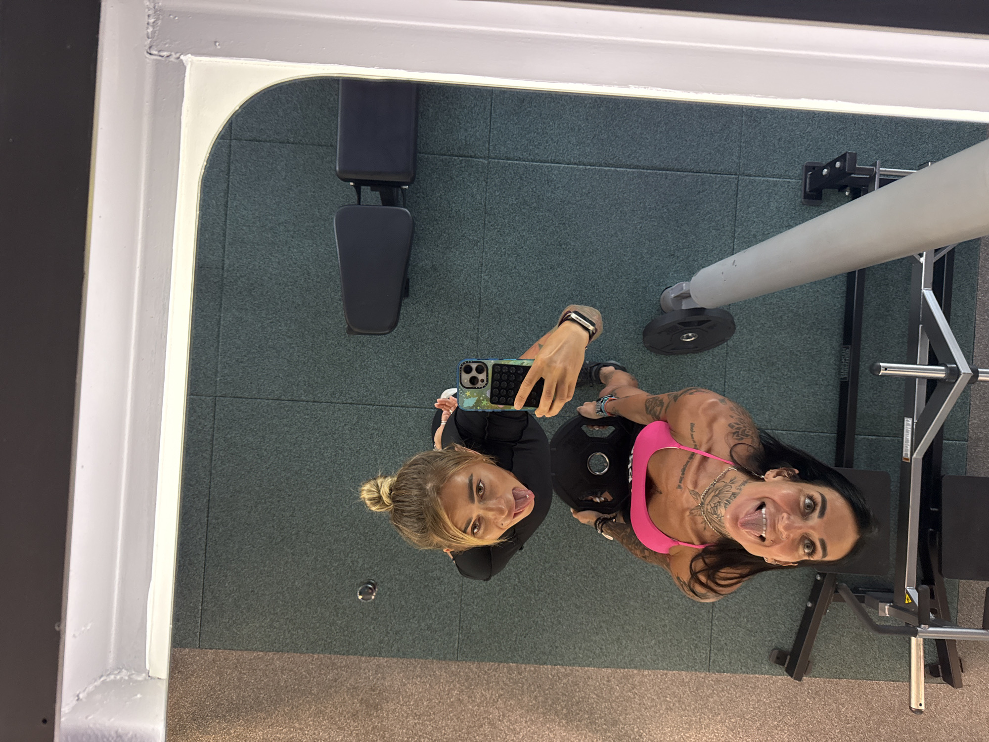 The image is a reflection in a gym mirror, showing two individuals. The person in the foreground is wearing a bright pink sports bra and has numerous tattoos. They are sticking their tongue out and looking at the camera, with their head tilted back. They are holding a weight plate. The second person, reflected behind them, is wearing a black top and has their tongue sticking out and a piercing in their lip. They are holding a smartphone to take the picture. The gym floor is covered in dark green mats. In the background, there are weight benches and gym equipment. The scene is an indoor gym environment.