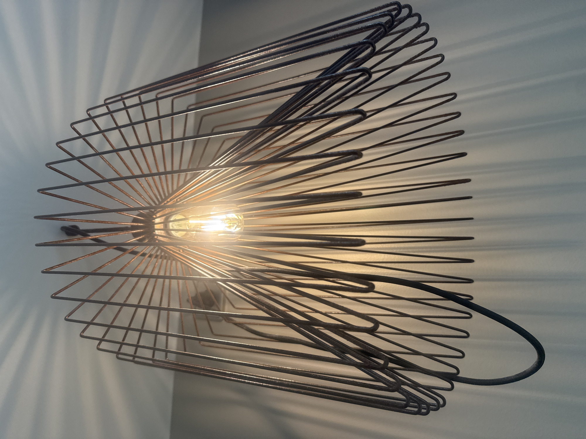 An interior shot of a modern, wire-frame pendant light fixture is captured from a low-angle perspective against a pale blue wall. The light fixture, made of thin, dark metal wires, has a wide, conical shape, with wires radiating outwards and downwards from a central point where a warm-toned Edison-style light bulb is illuminated. The light bulb casts a soft glow and creates a starburst pattern of shadows on the ceiling and upper wall. A black electrical cord is visible hanging from the side of the fixture. The overall impression is one of contemporary, minimalist design. The scene is located indoors, presumably in a residential or commercial space.