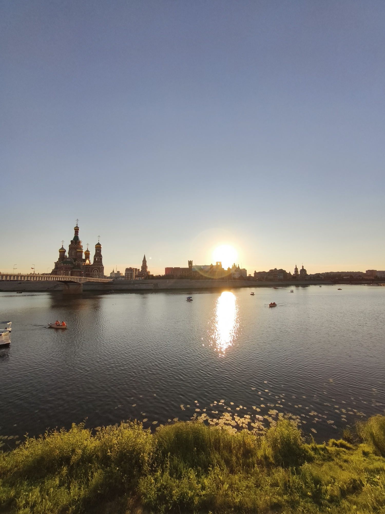 The image captures a sunset scene in Yoshkar-Ola, Russia, with the striking Orthodox cathedral as a prominent focal point. The bright, setting sun is positioned slightly to the right of the center, casting a warm, golden glow across the sky and reflecting brilliantly on the calm waters of the river.
In the foreground, the river's surface is rippled, and patches of water lilies are visible along the bank. The riverbank itself is covered in lush green grass and vegetation, illuminated by the late afternoon sun.
Across the river, the city skyline is visible. The most prominent structure is a large, ornate Orthodox cathedral with multiple golden domes and intricate architectural details, situated on the left side of the frame. To its right, a bridge spans the river, connecting the cathedral to the cityscape beyond. Other buildings, including a tall tower with a spire, are visible further down the skyline.
Several small boats are scattered across the river. On the left, a white boat is partially visible. Further into the river, a small rowboat with at least two people wearing orange life vests is being paddled. Other small boats, possibly paddle boats or kayaks, are also present, with some appearing to be occupied. The overall atmosphere is serene and picturesque, highlighting the beauty of the city at sunset.
