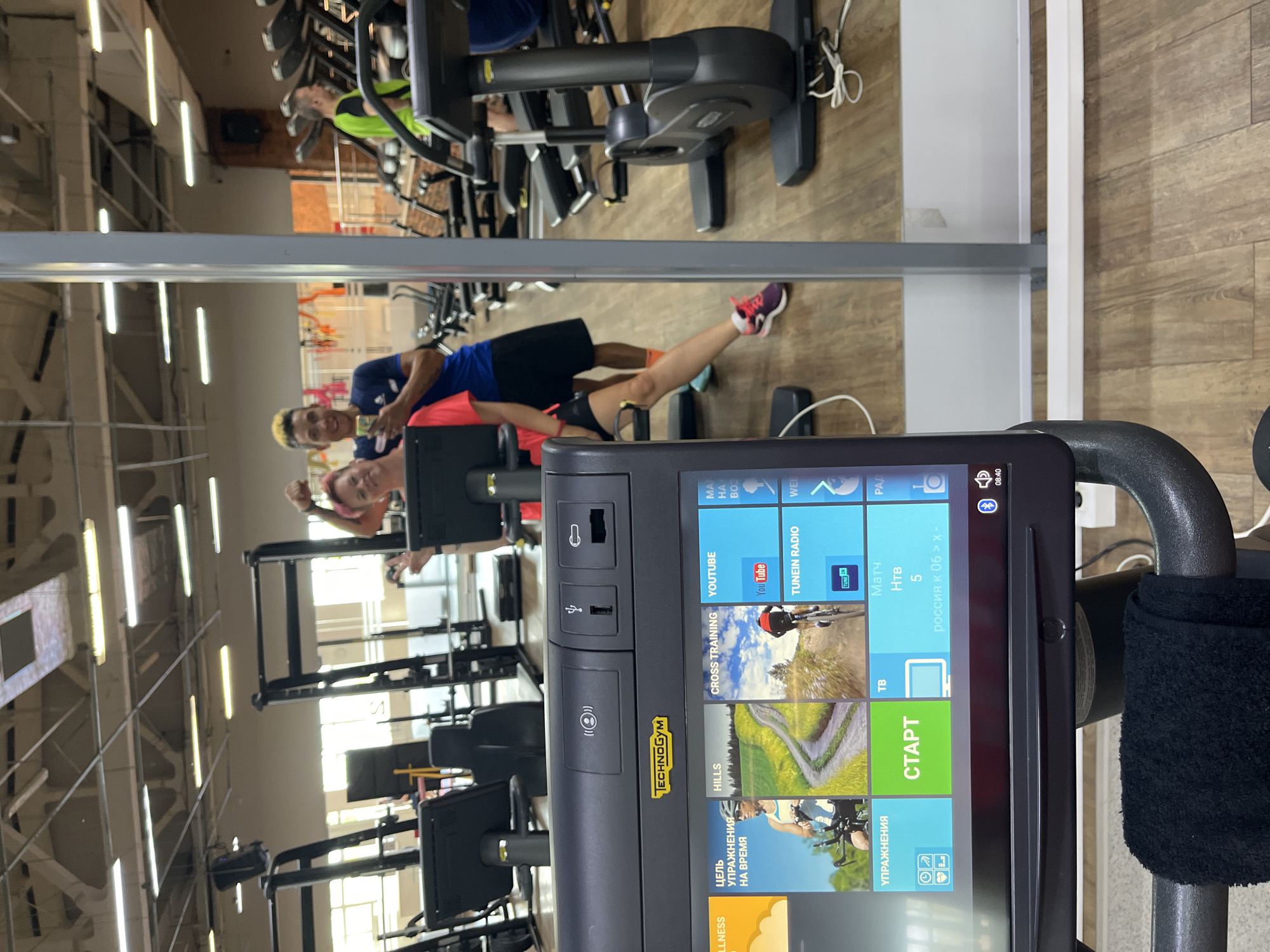 In a gym in the city of Chaikovsky, Russia, a woman with pink hair and a man with short dark hair are taking a selfie in front of a gym bike's monitor. The woman is wearing a pink t-shirt and black shorts, while the man is wearing a blue t-shirt and black shorts. The gym bike's monitor displays various workout options, including "Wellness," "Goals," "Hills," "Cross Training," and "YouTube." Other gym equipment, such as treadmills and weight machines, are visible in the background. The overall environment appears to be a modern and well-equipped fitness facility.